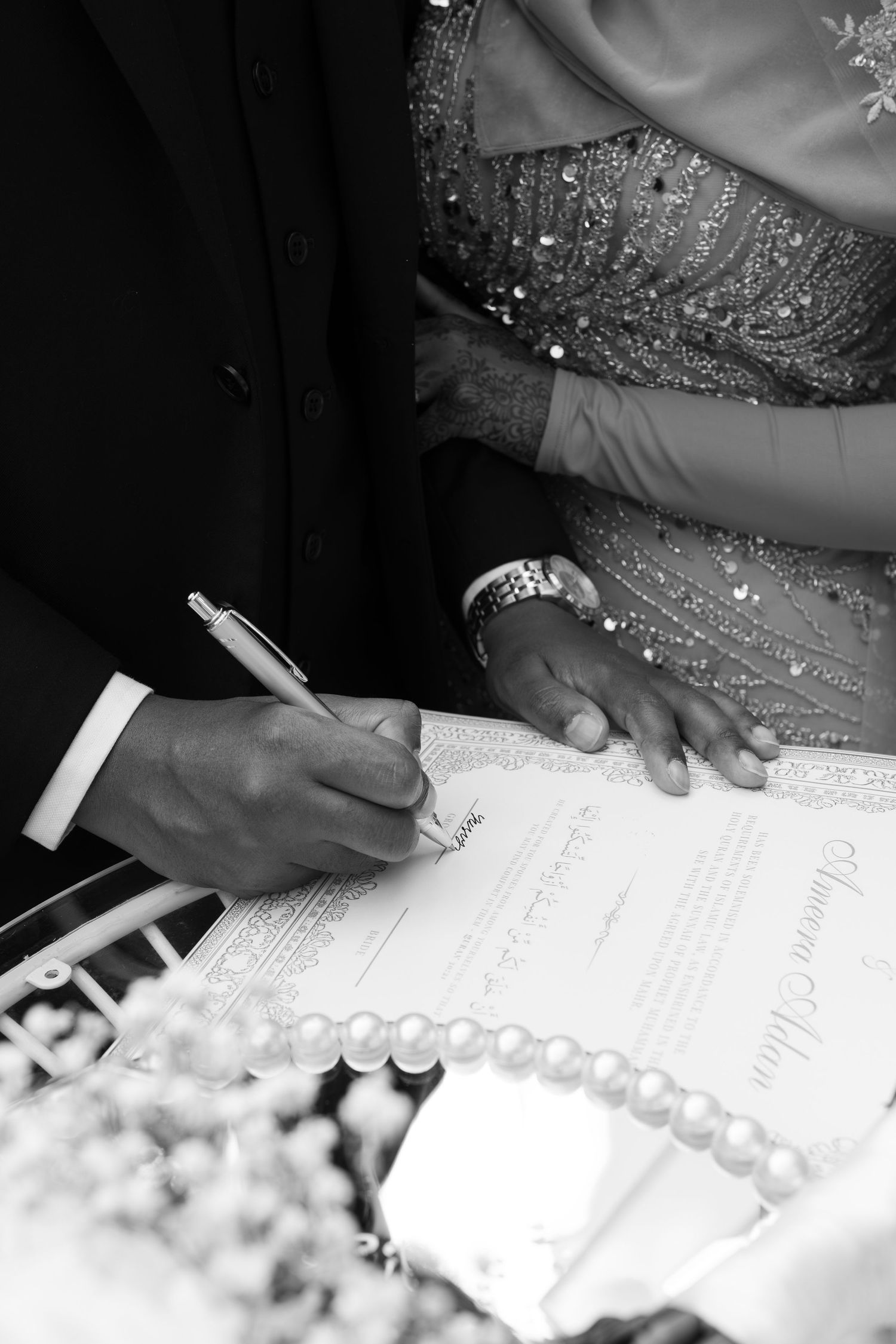 Black and white photo of couple signing wedding certificate, bride in beaded gown, pearl bouquet in foreground.