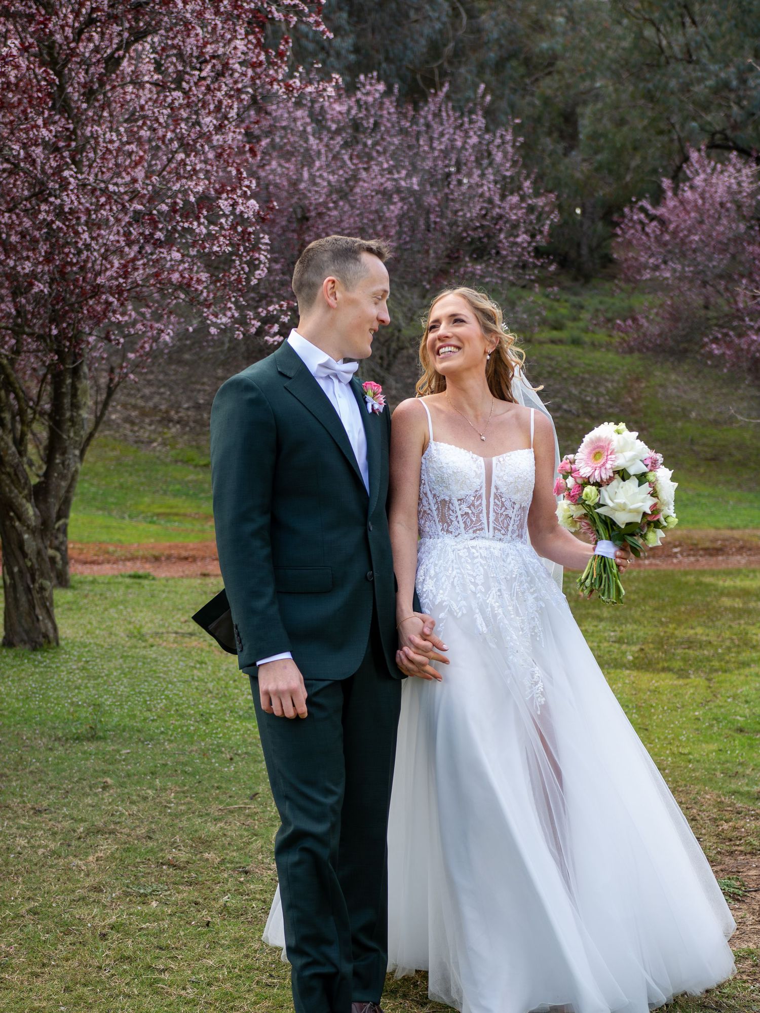 Bride and groom holding hands in front of blooming pink cherry blossom trees, bride holding a bouquet.