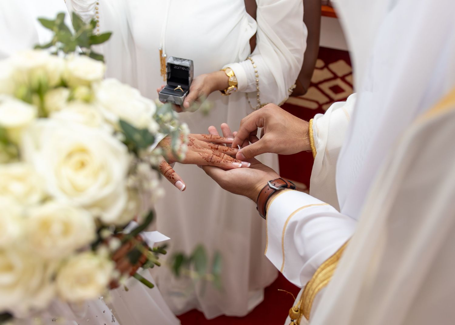 A wedding ring exchange ceremony with white roses, gold jewelry, and elegant white attire in a formal setting.