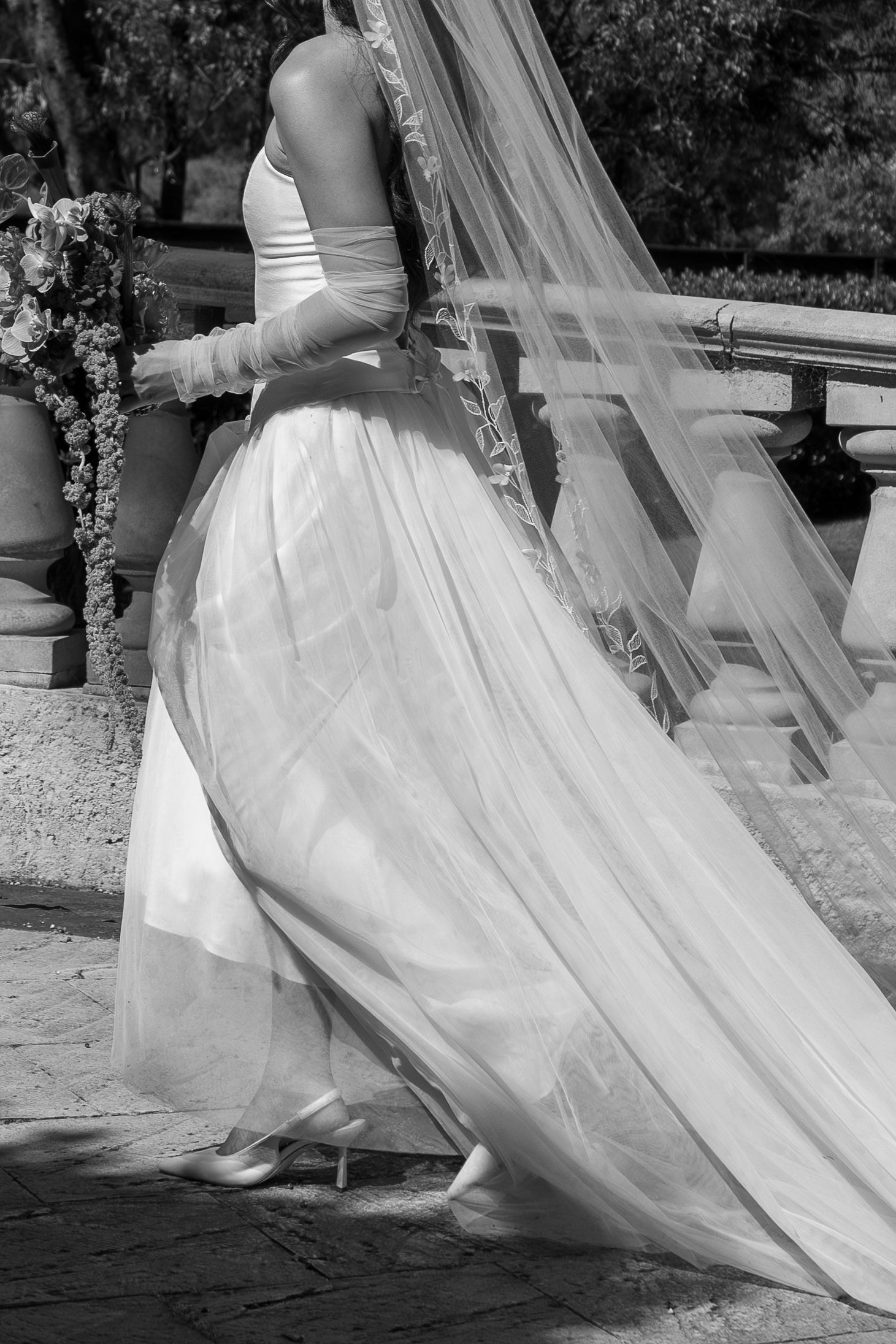 Black and white photo of a bride in a flowing white gown and veil walking on a stone terrace.