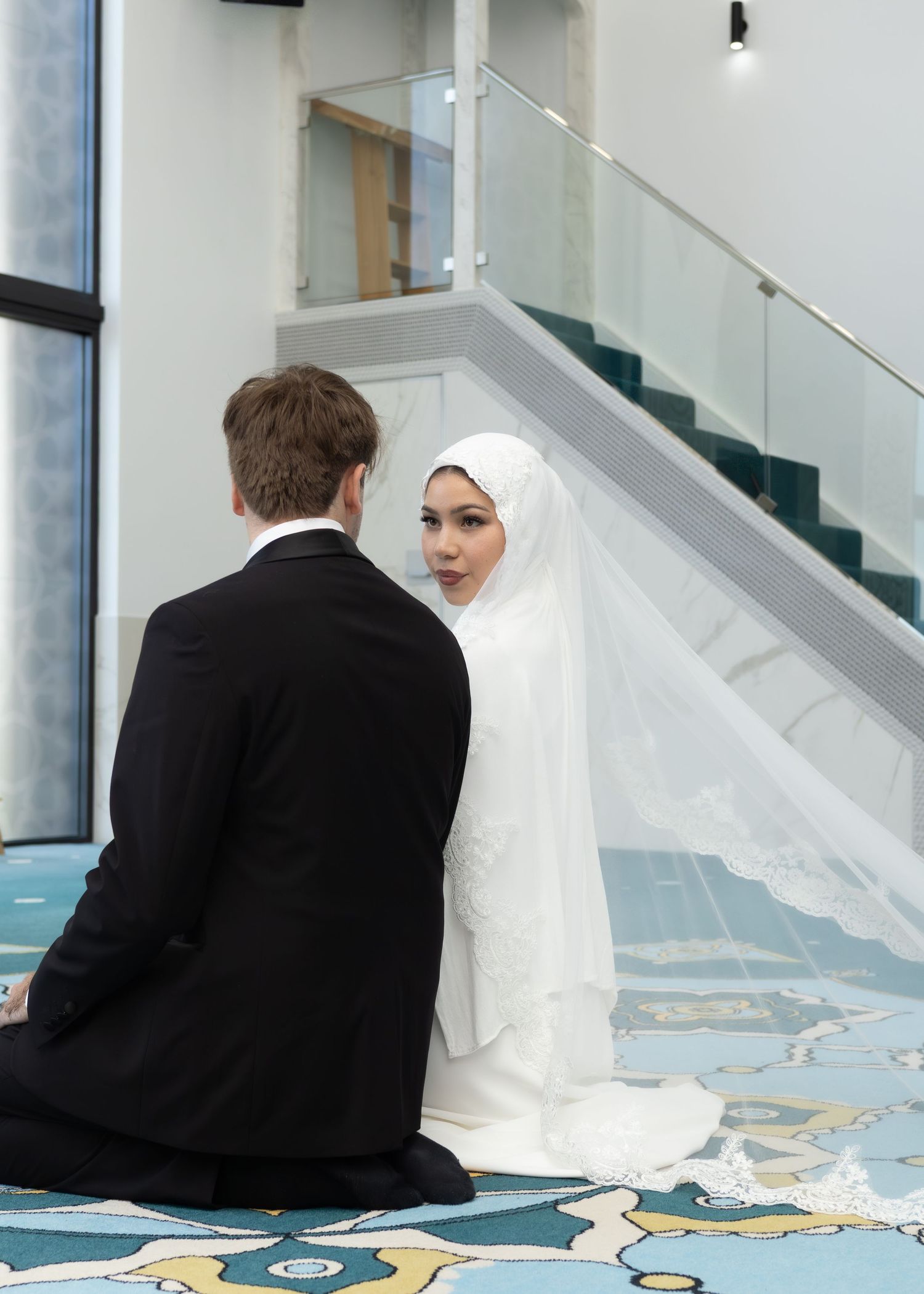 Muslim bride in white hijab and gown sits with groom in black suit during nikah wedding ceremony indoors.