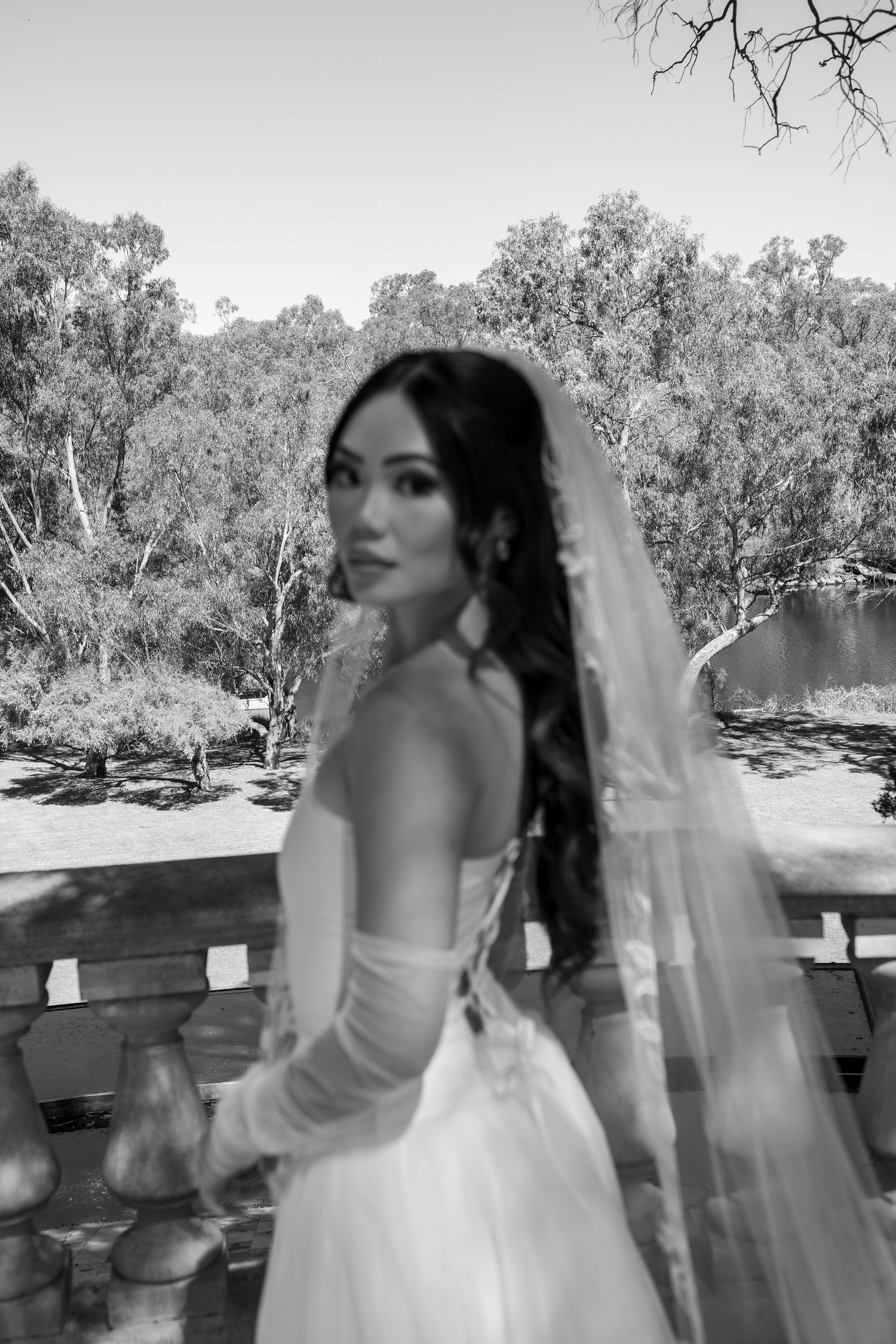 Black and white photo of a bride in a white gown and veil, standing on a balcony with trees in the background.