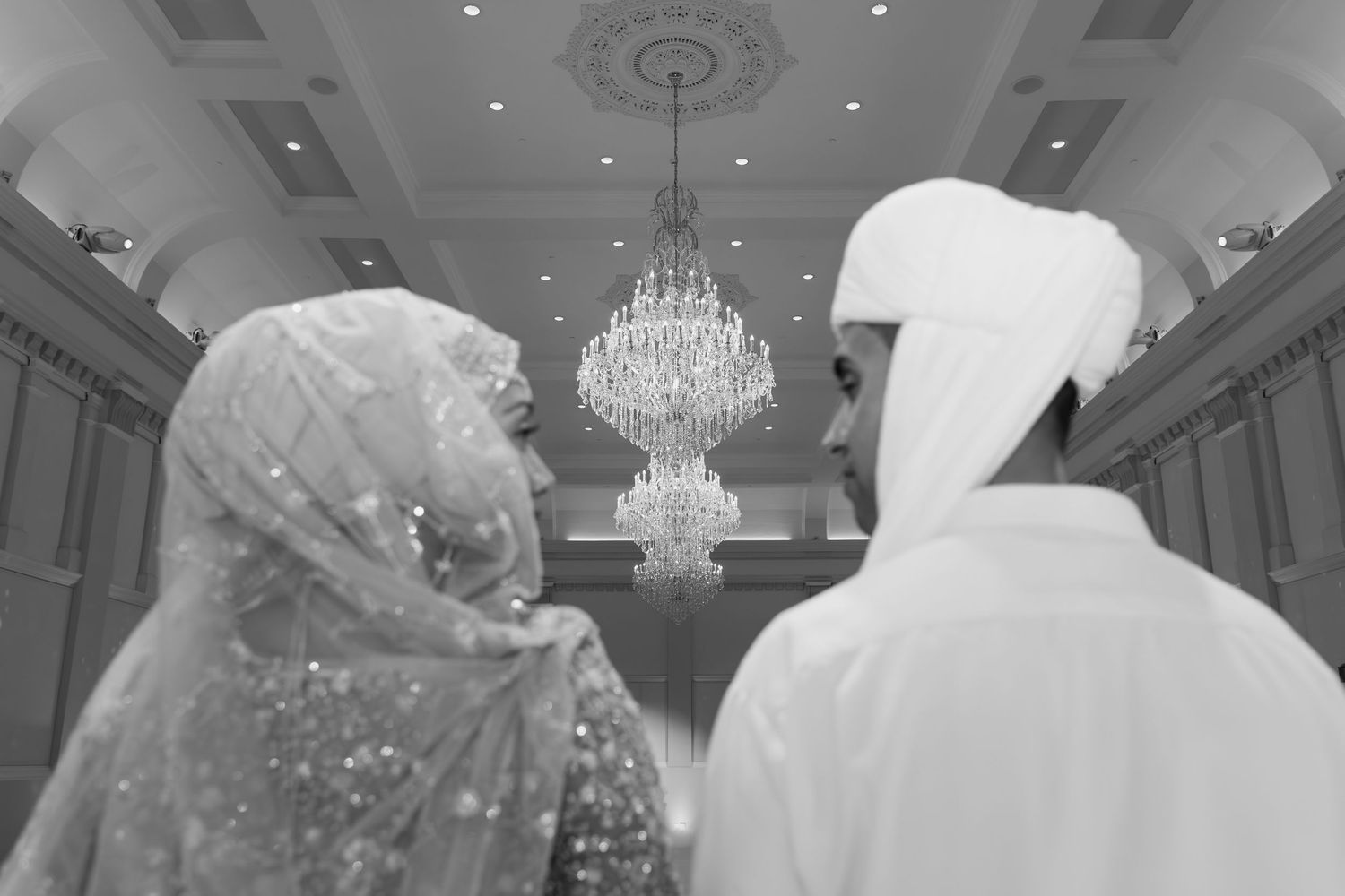 Black and white photo of Muslim bride and groom in elegant hall with crystal chandelier at their wedding.