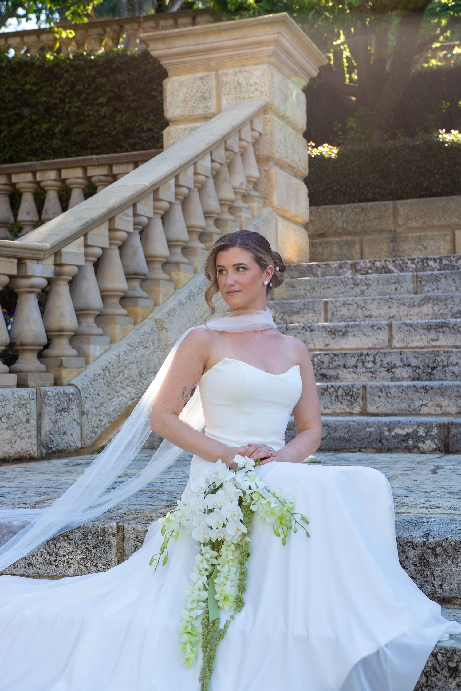 Bride in strapless white gown sits on stone steps holding cascading white floral bouquet with veil flowing behind her.