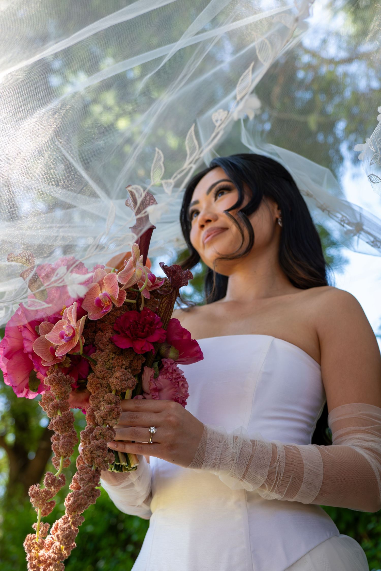 Bride in white strapless gown holding pink floral bouquet under veil outdoors.