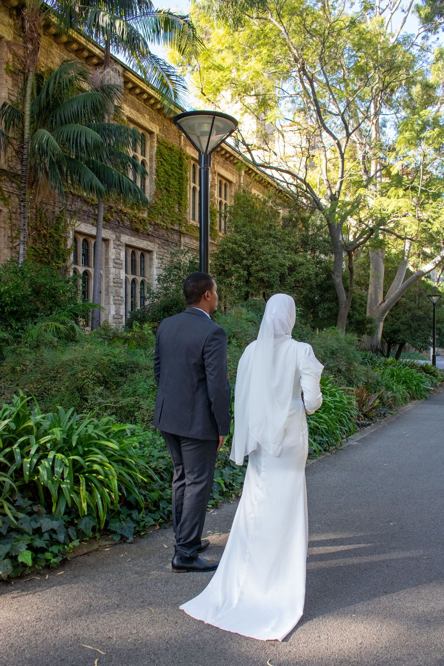 A bride in white hijab and groom in gray suit walk hand in hand along a garden path near an ivy-covered building.