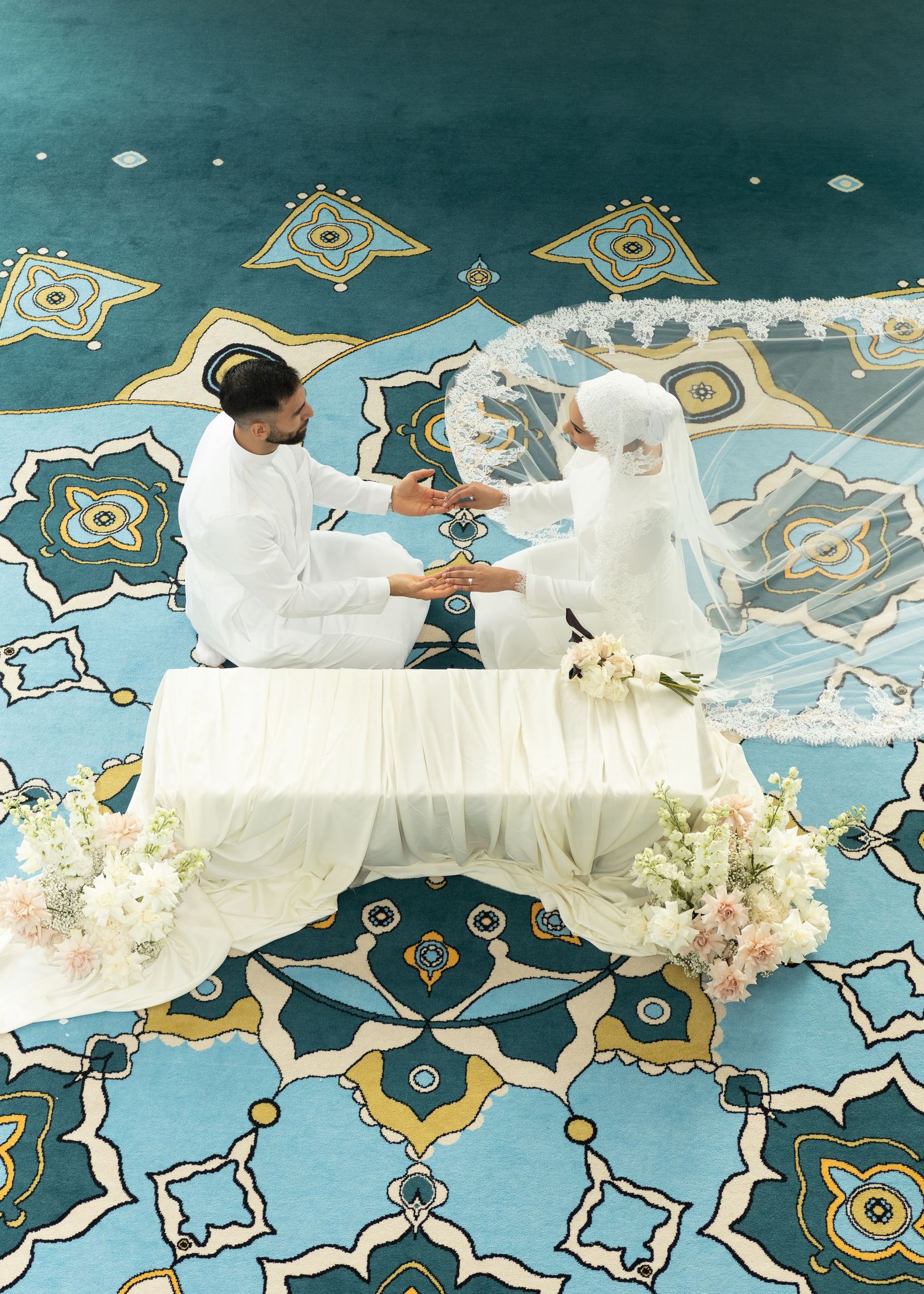 Muslim couple in white wedding attire seated on decorated platform with floral arrangements on ornate blue carpet.