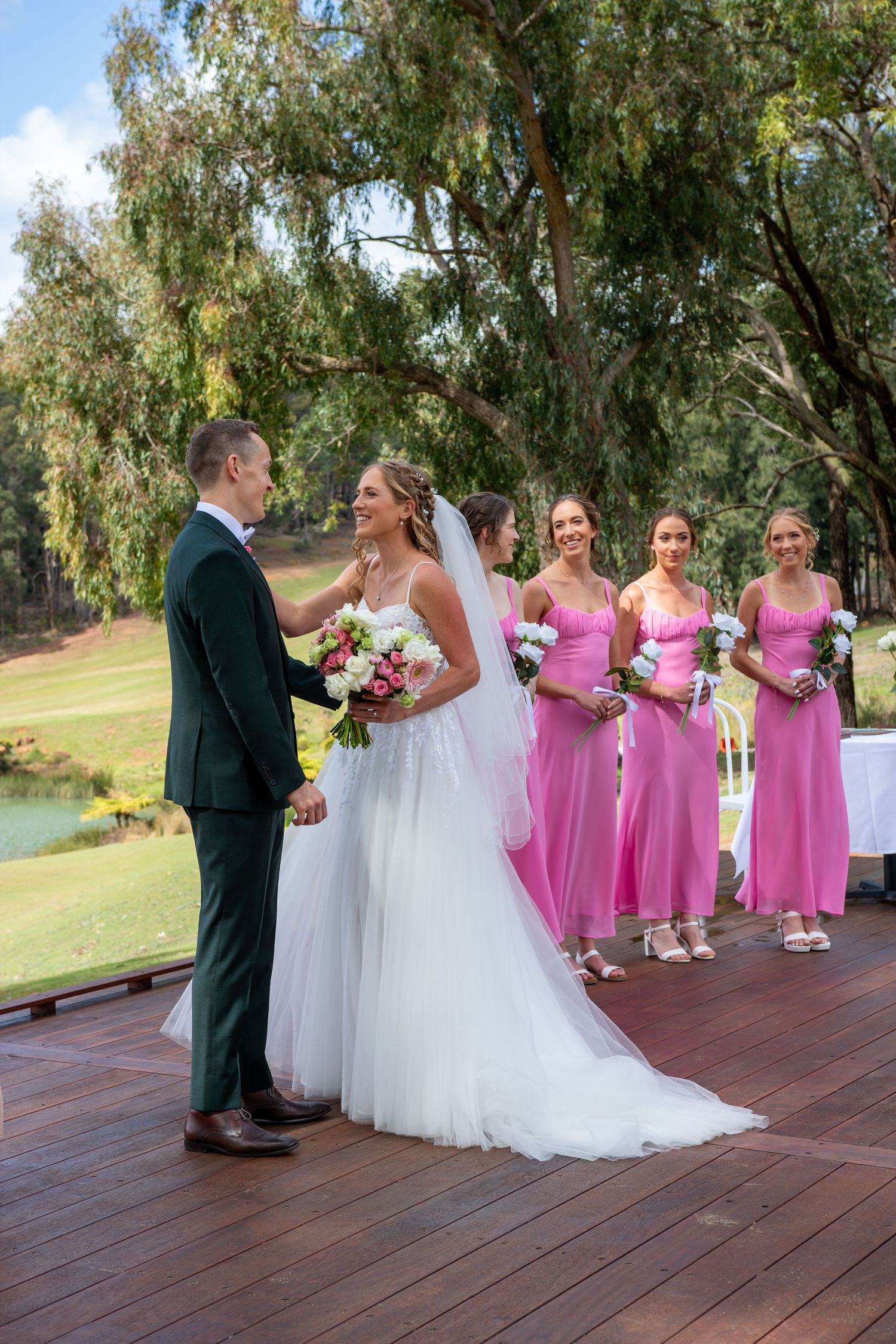 Bride and groom hold hands at outdoor wedding ceremony with four bridesmaids in pink dresses on wooden deck.