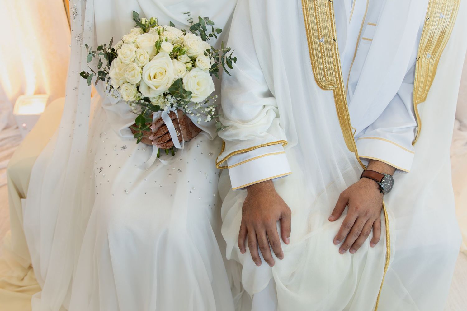 Bride and groom in white traditional wedding attire, bride holding white rose bouquet, close-up of hands and outfits.