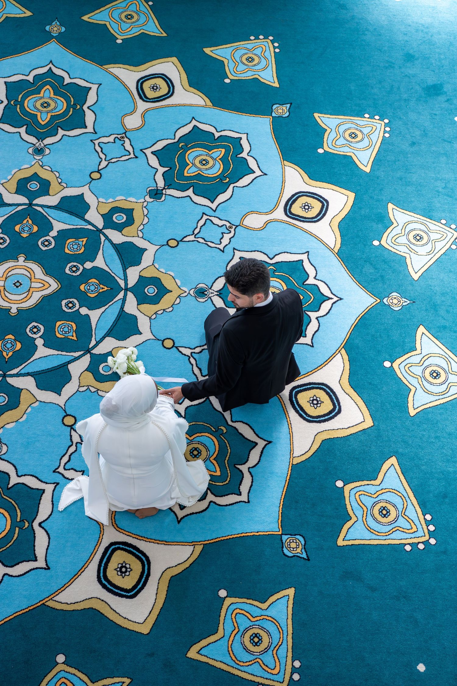 Aerial view of a couple in wedding attire standing on an ornate blue geometric patterned carpet in a mosque.
