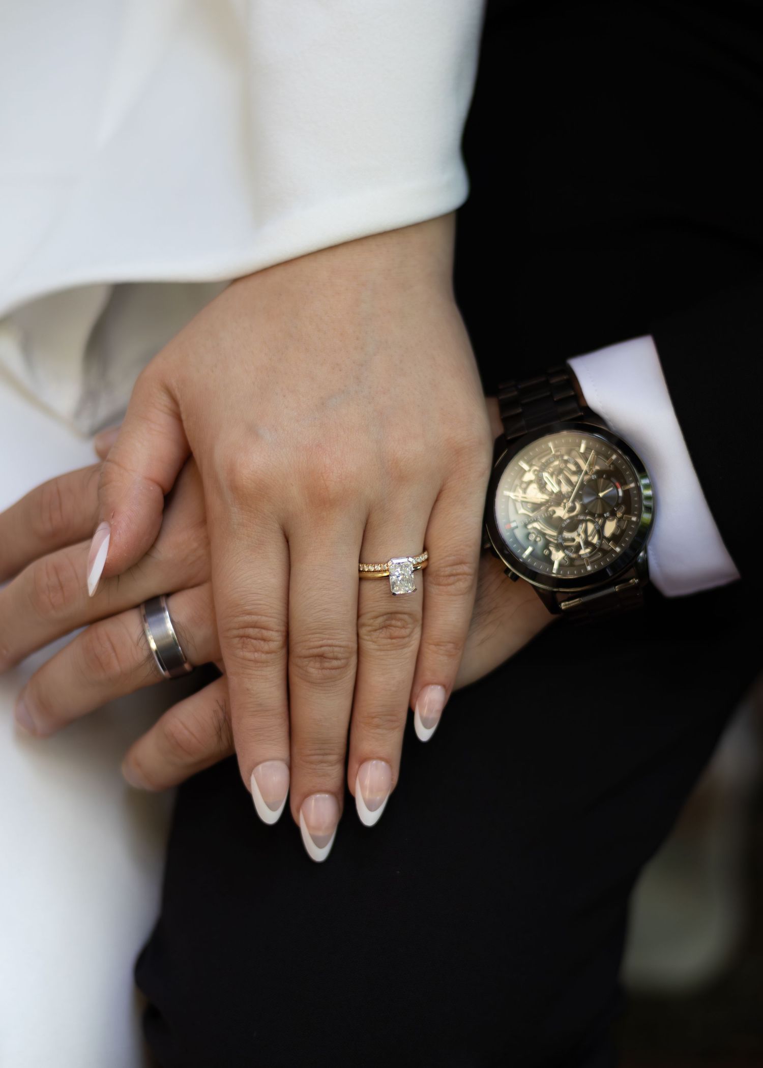 Close-up of newlywed couple's hands showing diamond engagement ring, gold wedding band, and black skeleton watch.