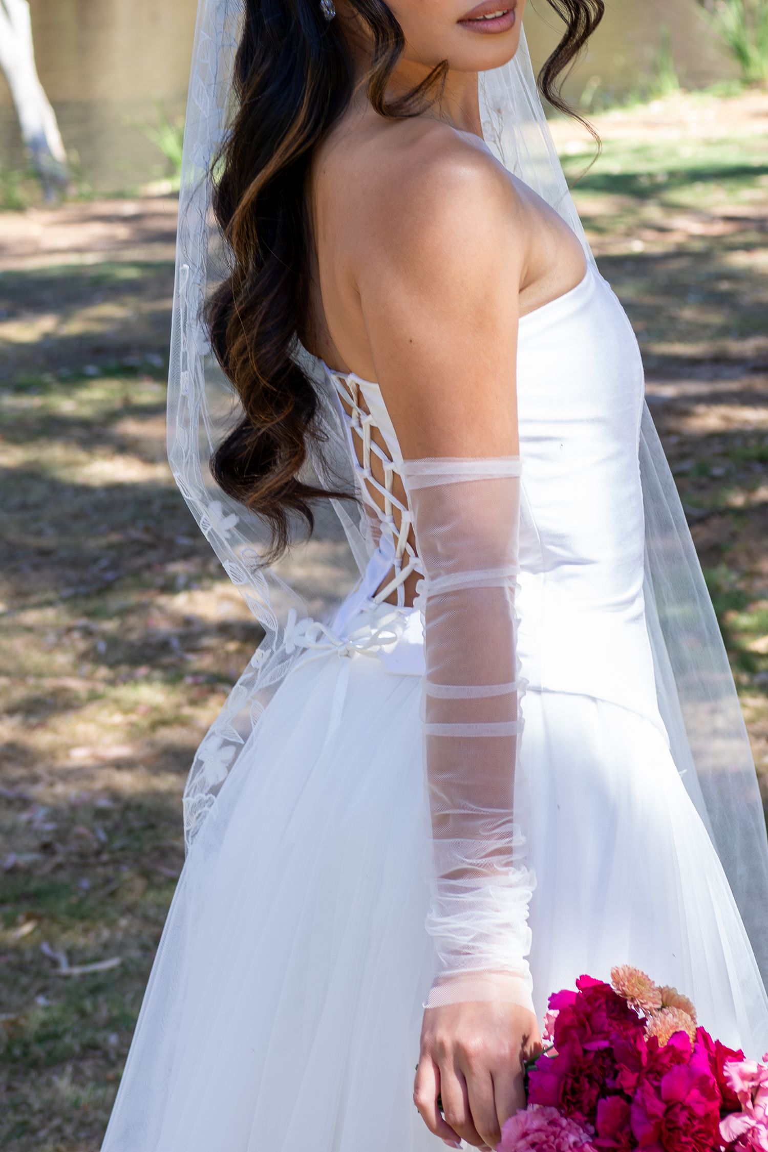 Bride in white strapless lace-up back wedding gown with sheer sleeves, veil, and pink bouquet outdoors.