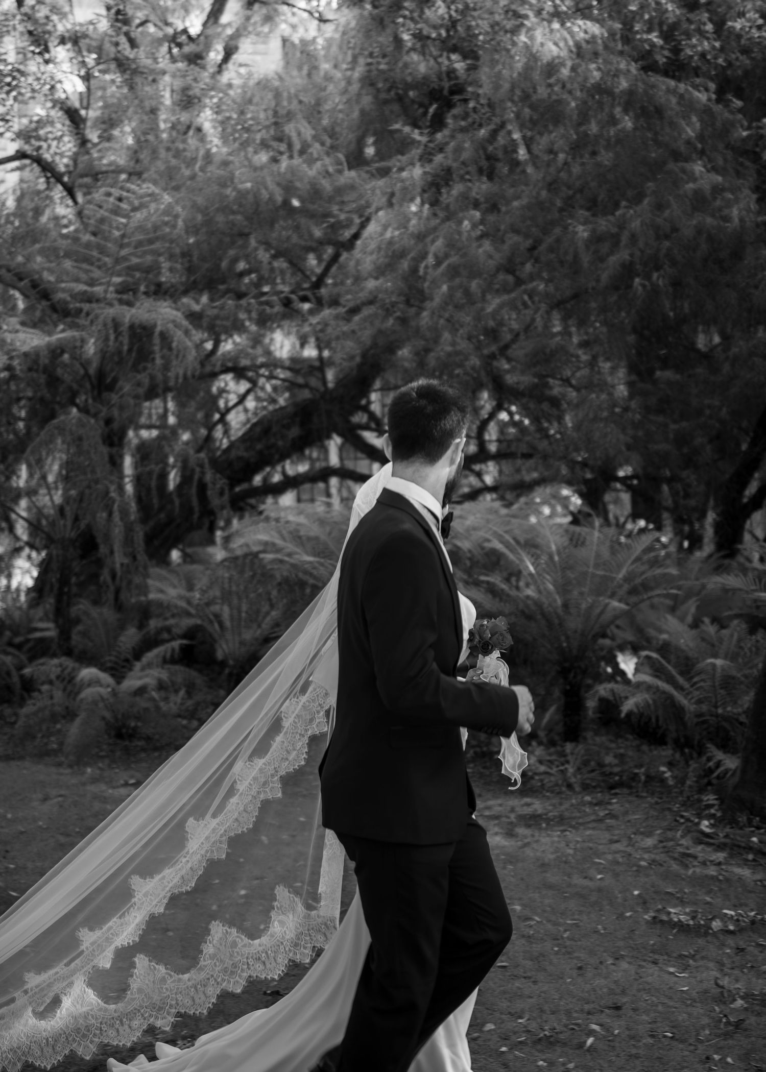 Black and white photo of a groom in a suit with a bride's lace veil flowing behind them in a lush garden.