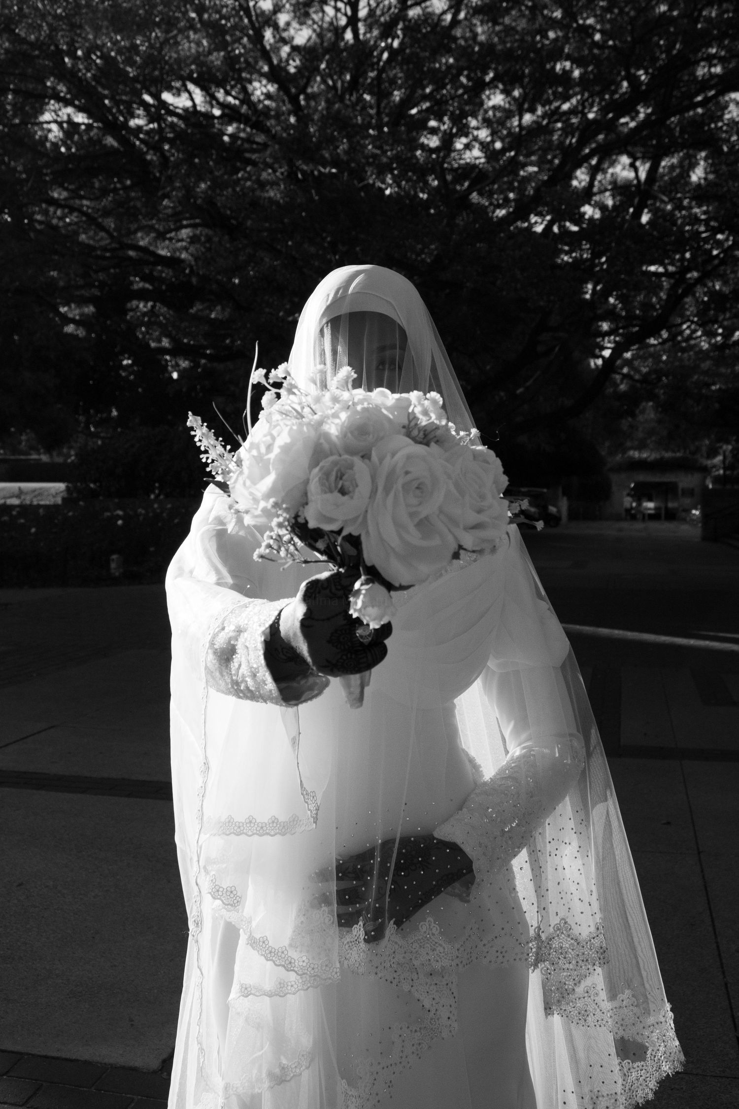 Black and white photo of a Muslim bride in white hijab and gown holding a bouquet of roses outdoors.