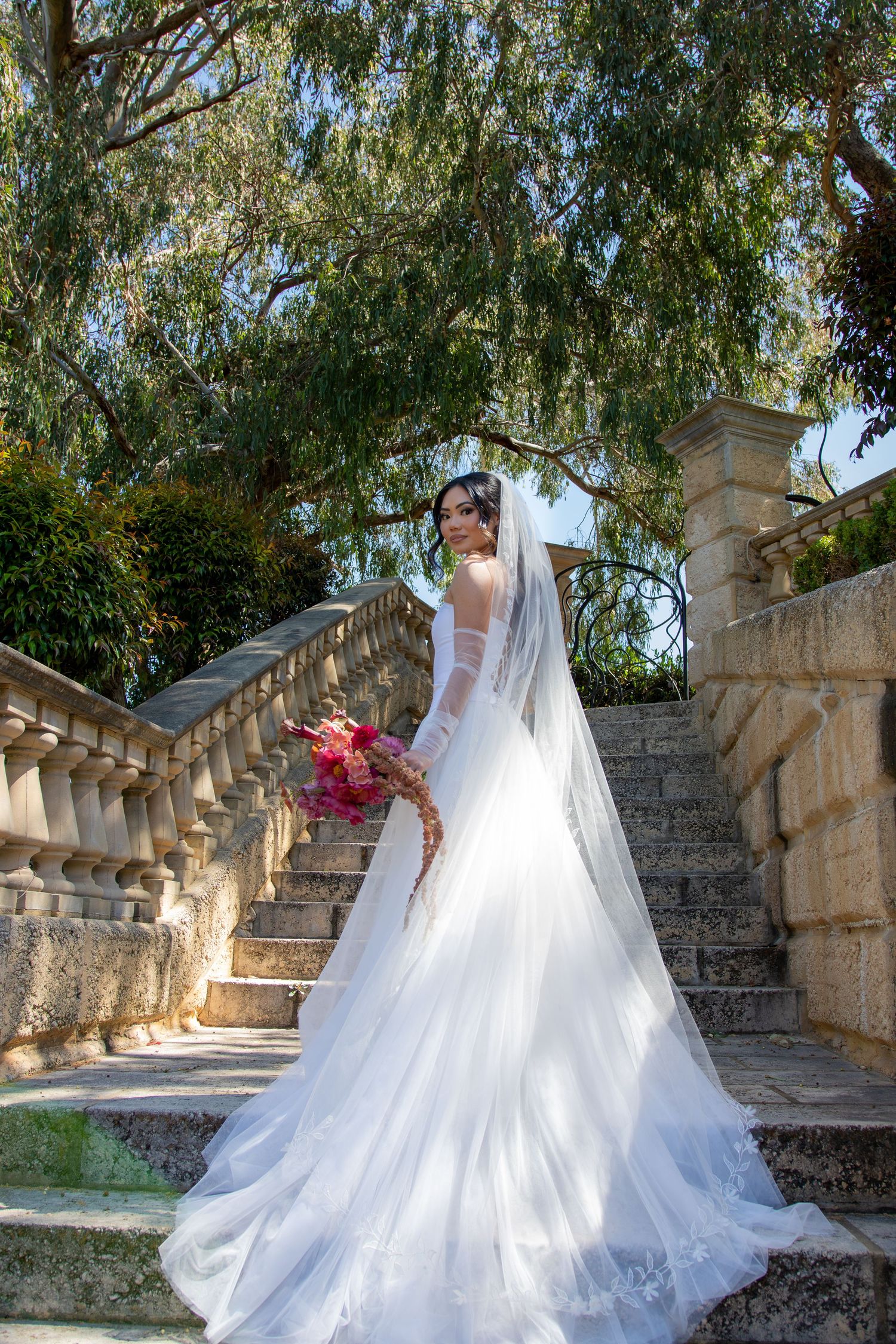Bride in white gown and veil holds red bouquet on stone steps surrounded by lush green trees.