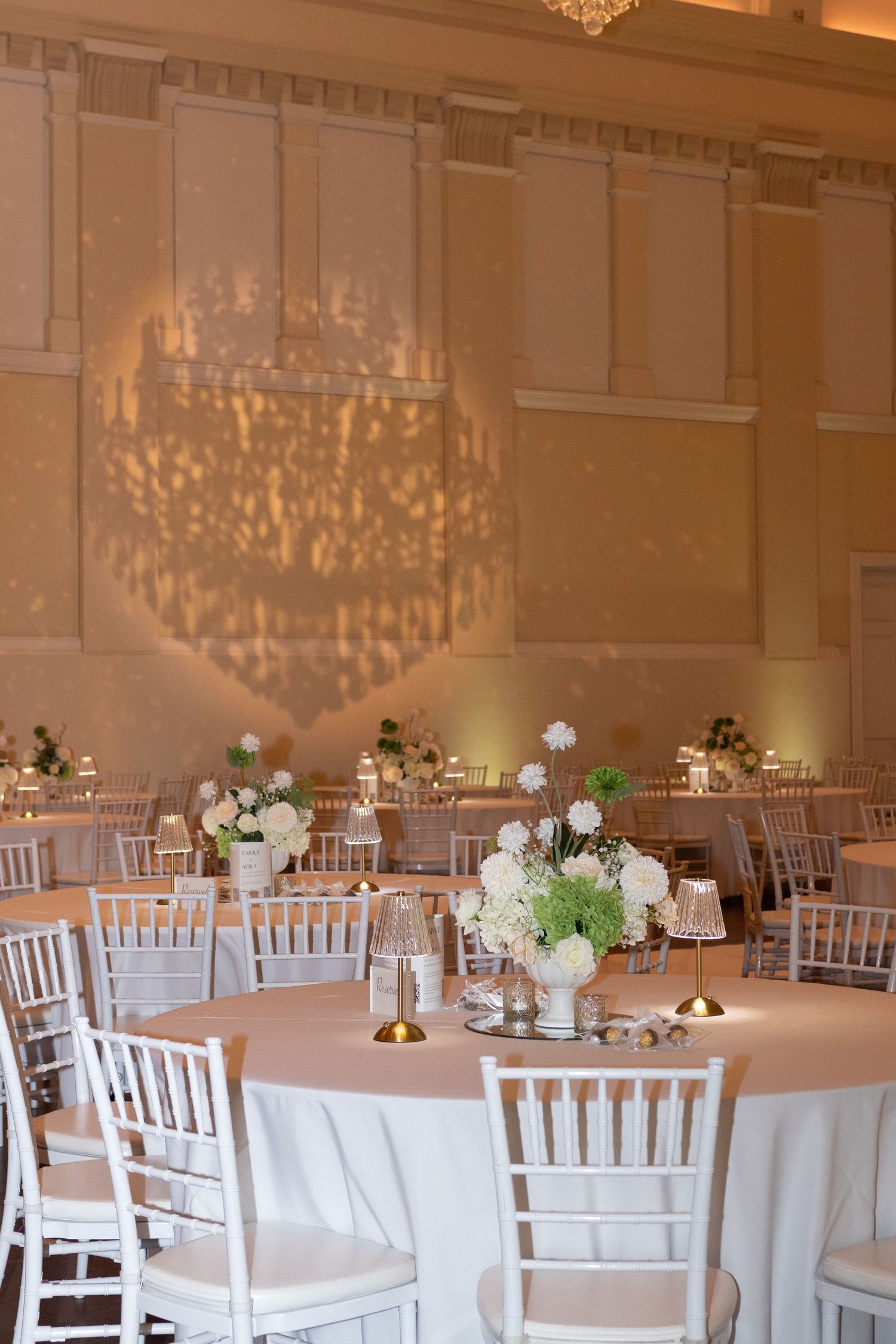 Elegant wedding reception ballroom with white Chiavari chairs, floral centerpieces, and warm chandelier light patterns on walls.