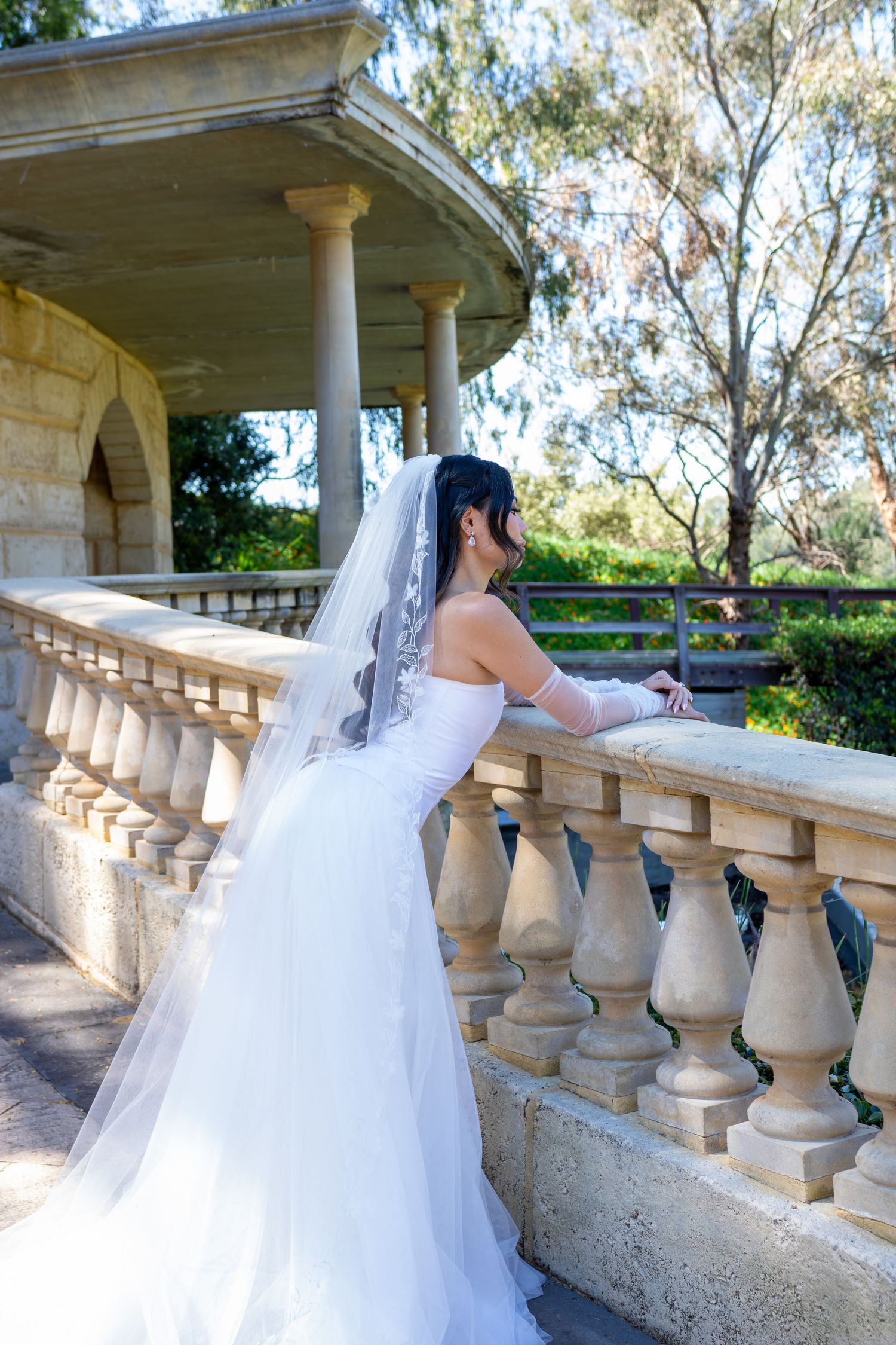 Bride in white gown and veil stands at stone balustrade balcony overlooking lush garden at elegant outdoor venue.