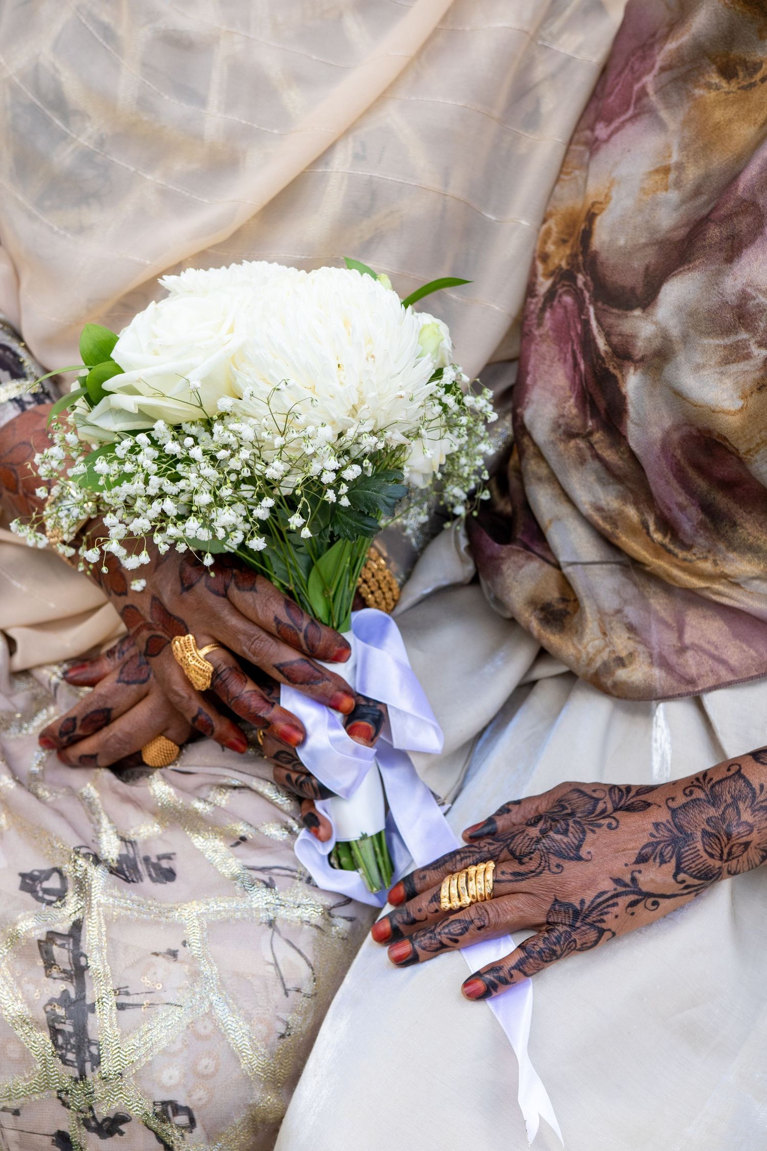Bride's hands with henna mehndi and gold rings holding white rose bouquet with ribbon on wedding dress.