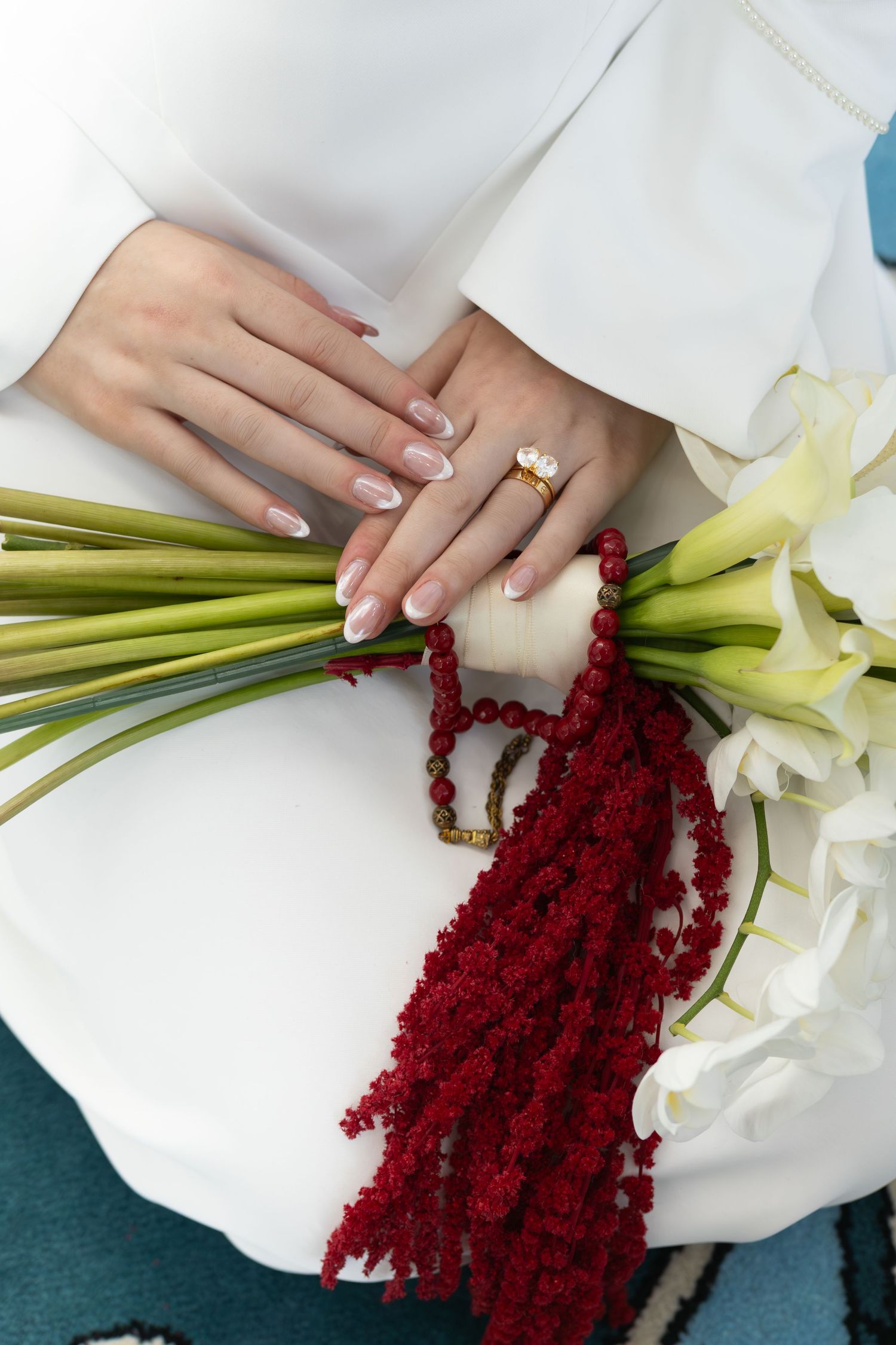 Bride holding white calla lily bouquet with red amaranthus, wearing gold ring and French manicure nails.