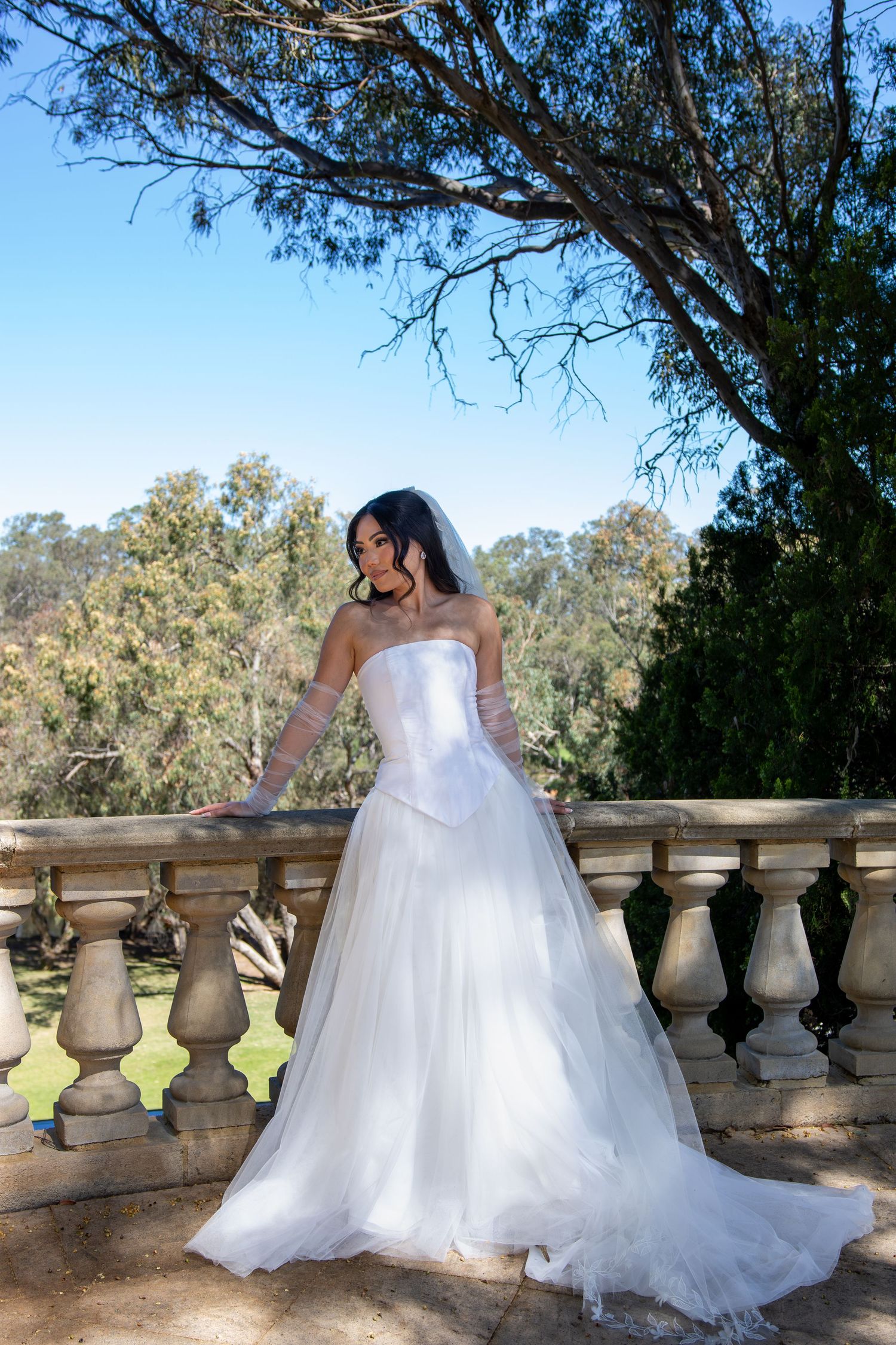 Bride in strapless white ballgown with tulle skirt poses on stone balustrade balcony surrounded by lush green trees.