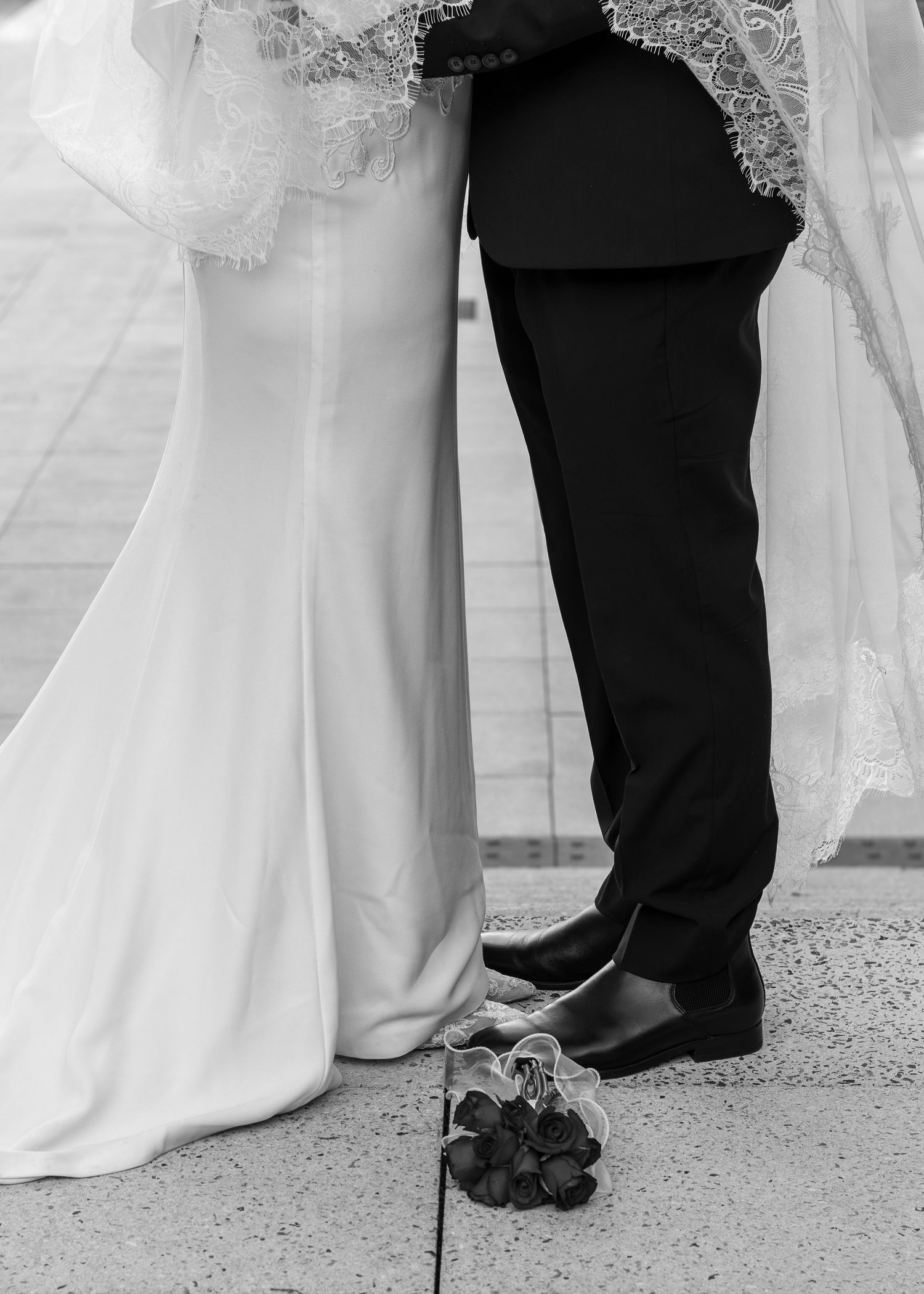 Black and white photo of bride and groom standing close, wedding dress and suit visible with floral bouquet on ground.