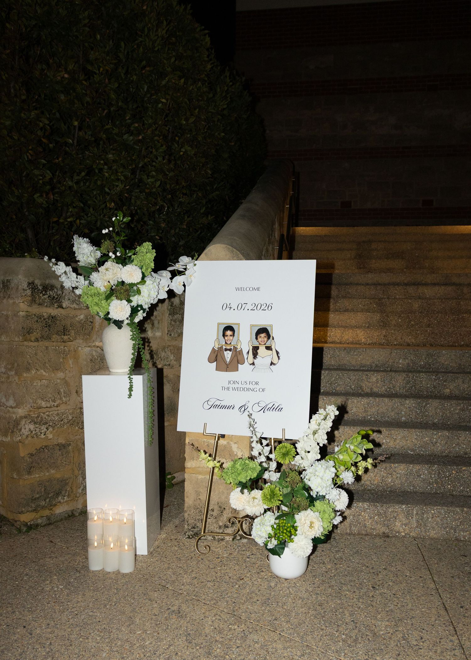 Elegant wedding welcome sign on easel with white floral arrangements and candles on stone steps at night.