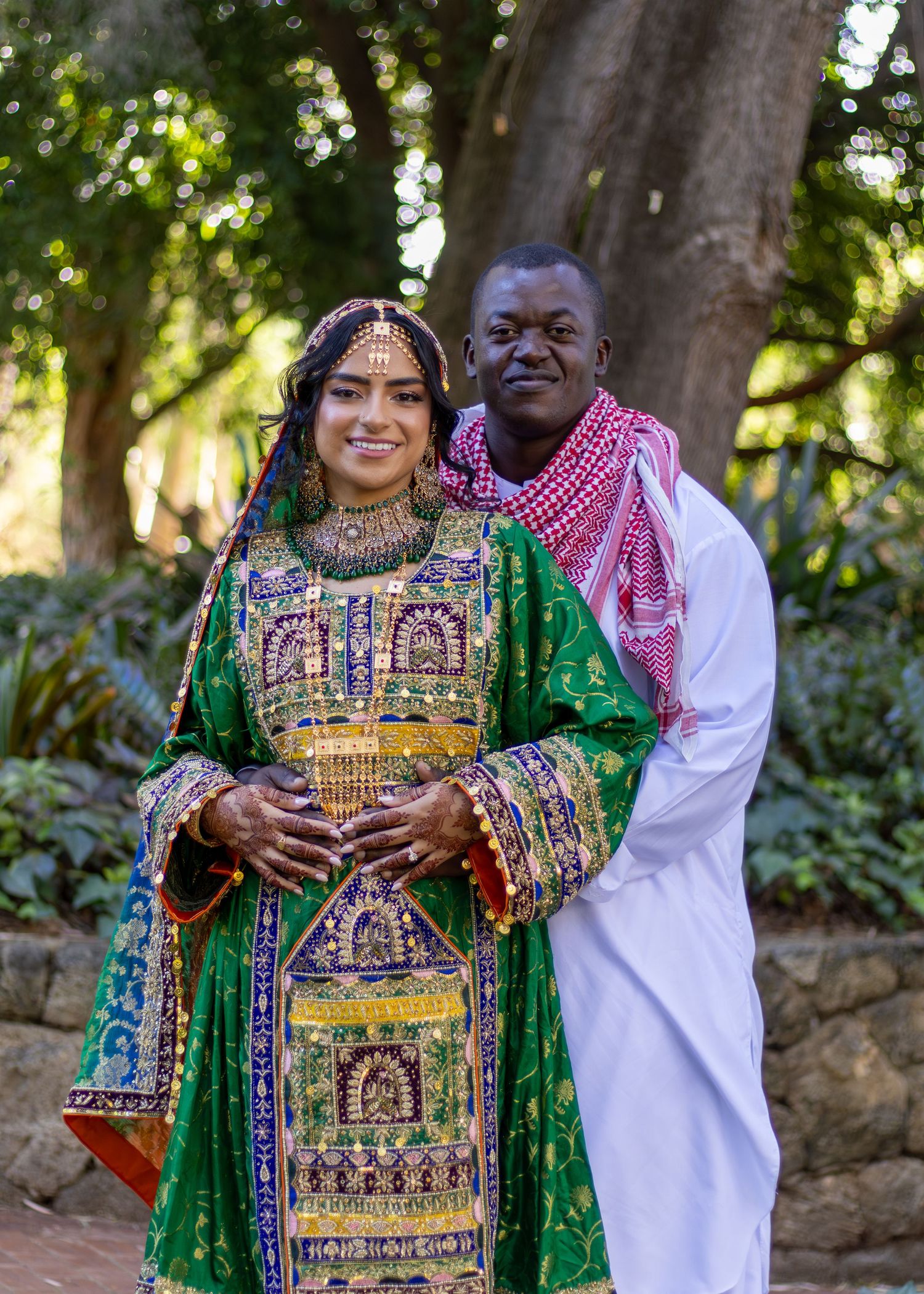 Couple in traditional cultural attire posing outdoors, woman in ornate green Afghan dress with gold embroidery and man in white thobe.