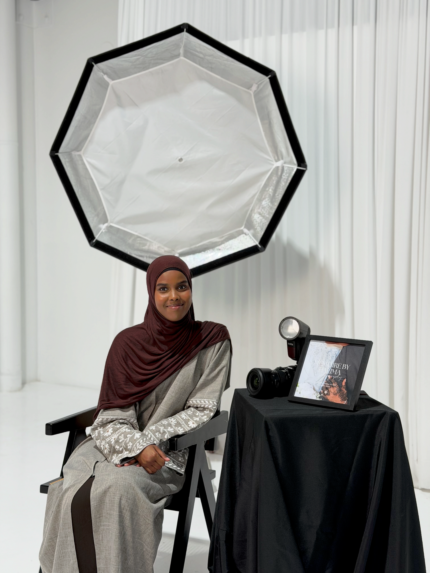 Woman in brown hijab and gray abaya sits before a large octagonal softbox in a bright photography studio.