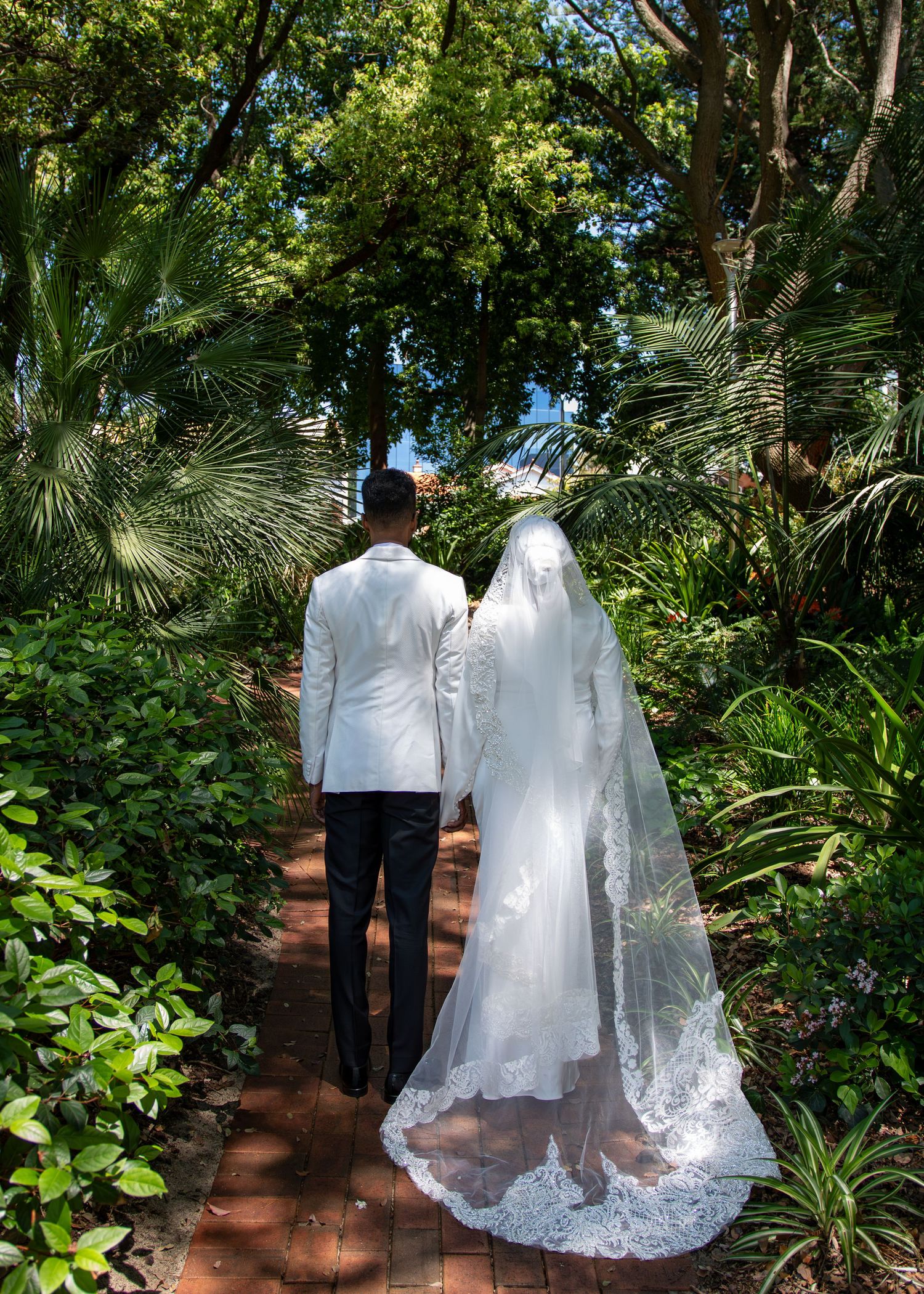 Bride and groom walking hand in hand down a brick garden path surrounded by lush tropical greenery on their wedding day.