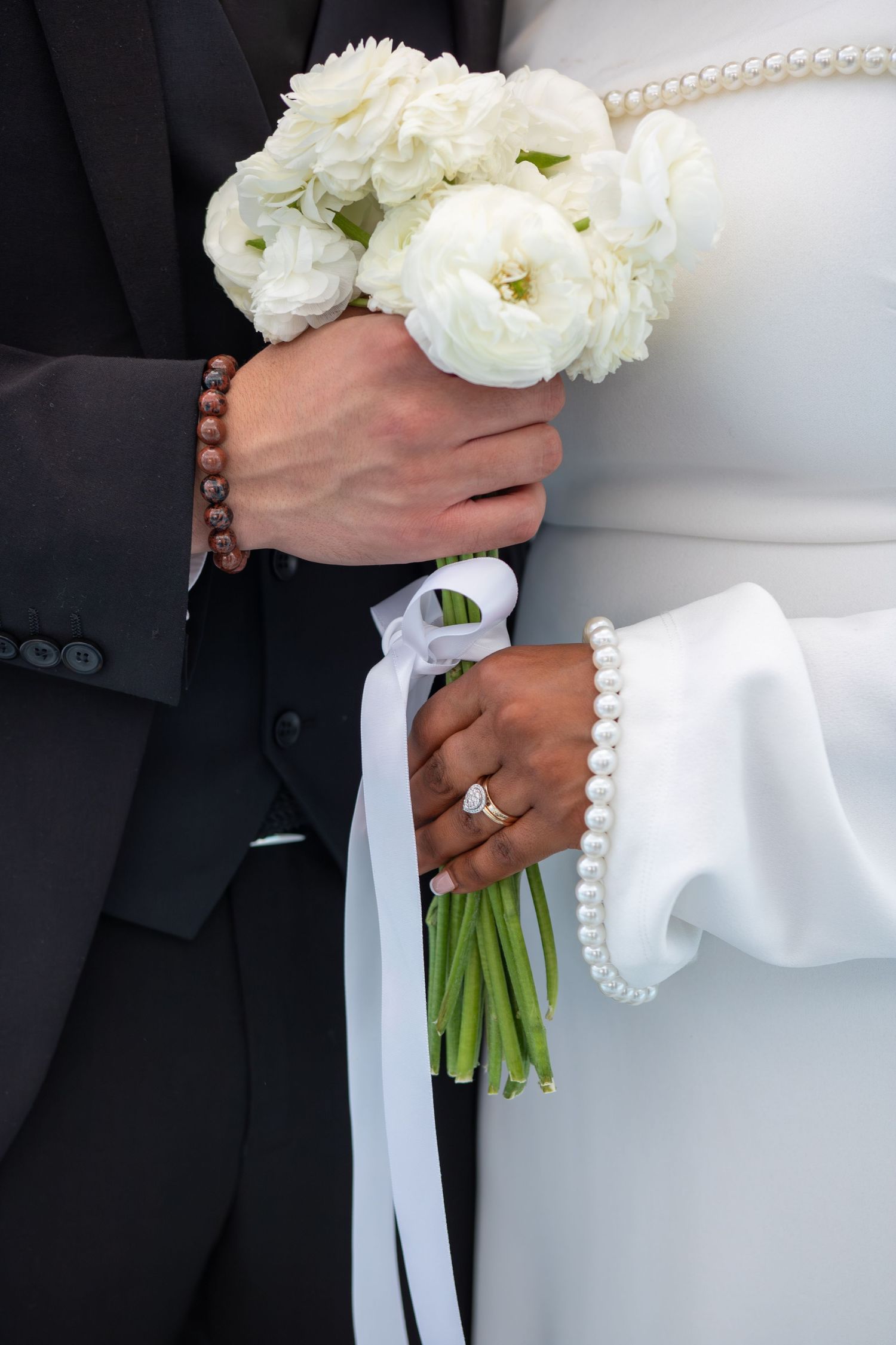 Bride and groom holding white ranunculus bouquet with ribbon, bride wearing diamond ring and pearl bracelet.
