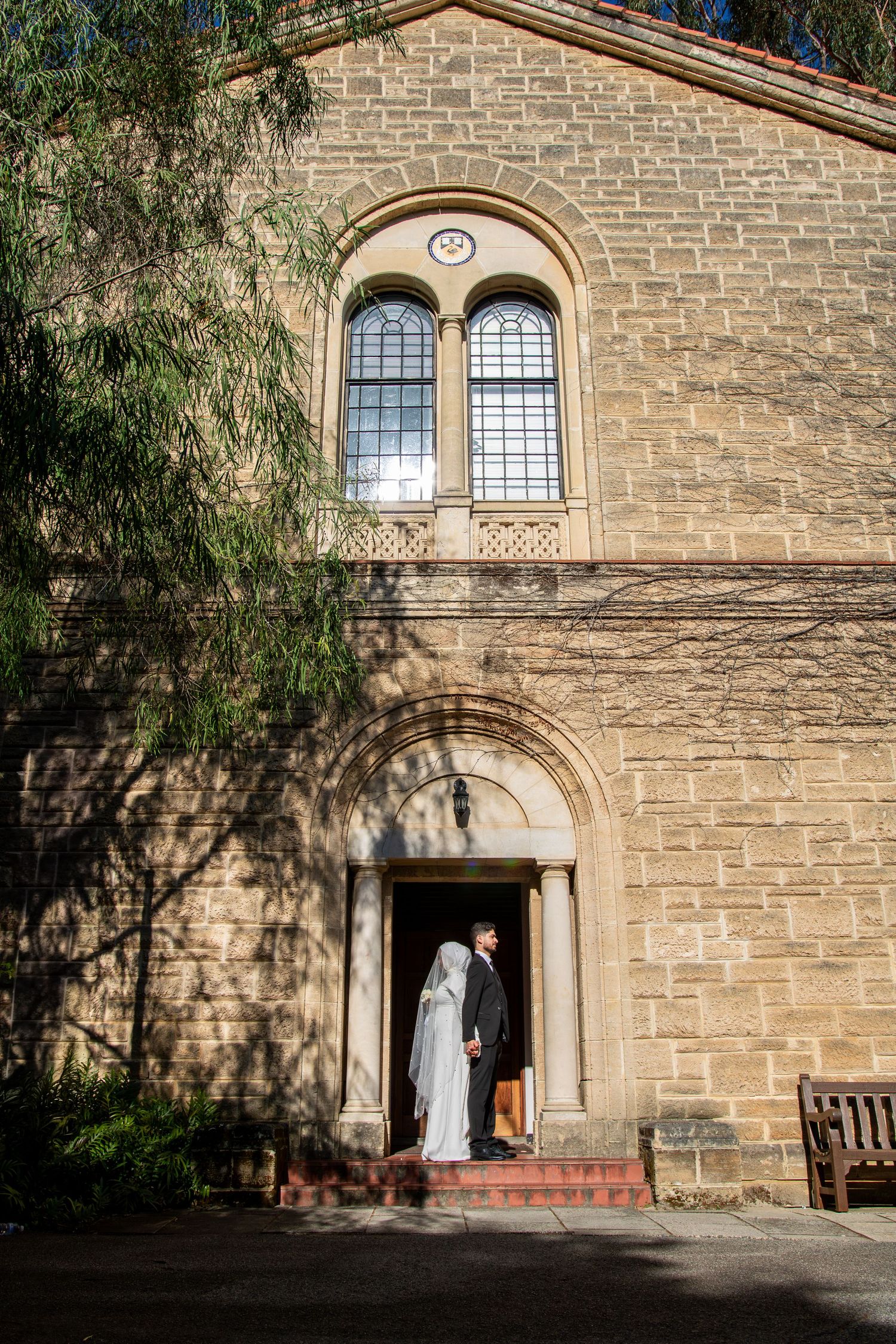 Bride and groom standing in arched doorway of historic sandstone church building with elegant windows.
