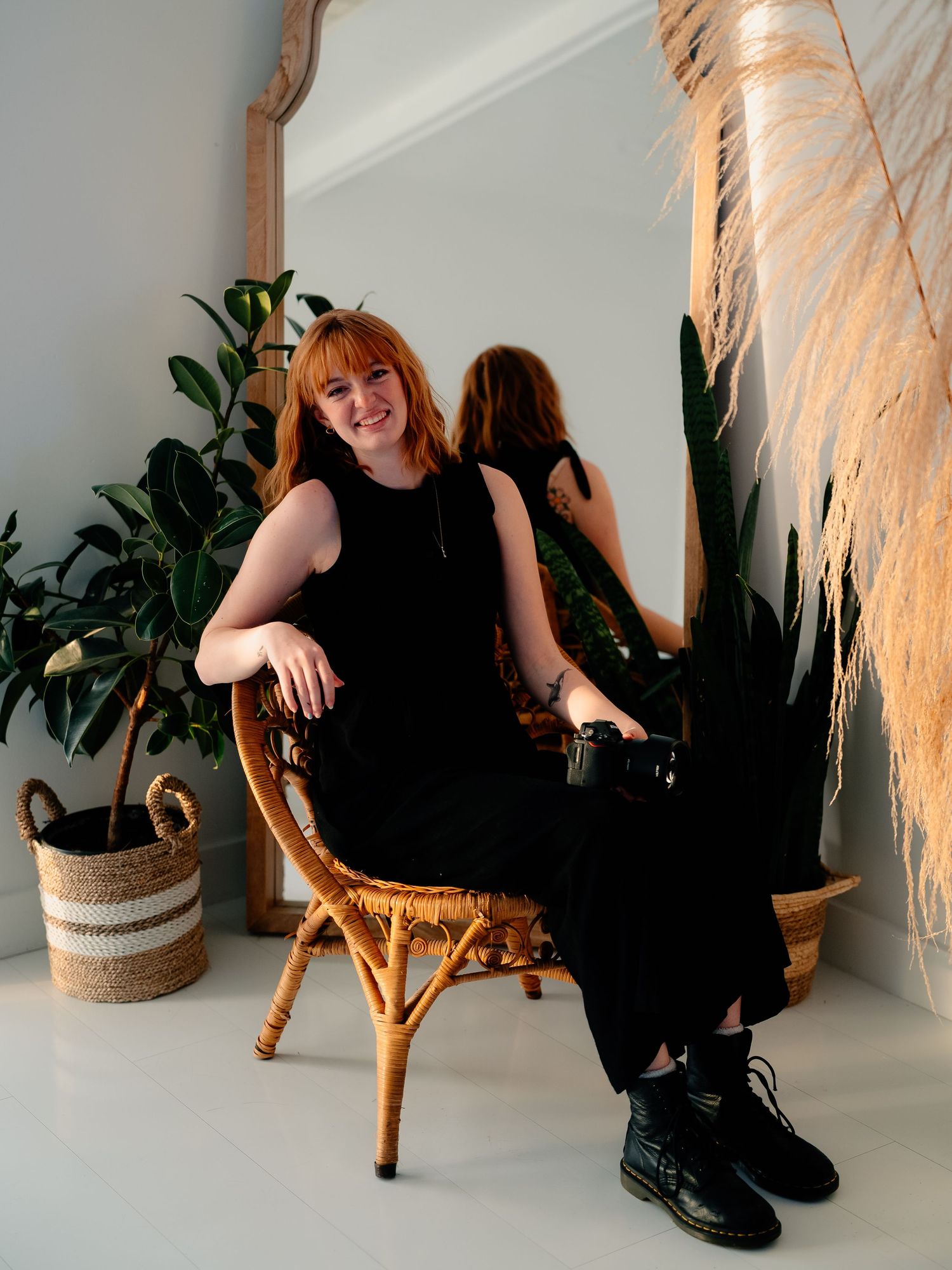 A person in a black outfit sits in a rattan chair next to a potted plant and mirror in a bright minimalist setting.