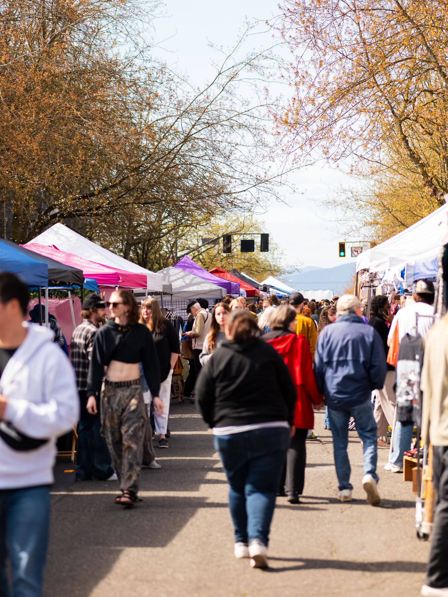 A panoramic view of a busy outdoor market with white tents and crowds browsing vendor stalls on an autumn day.
