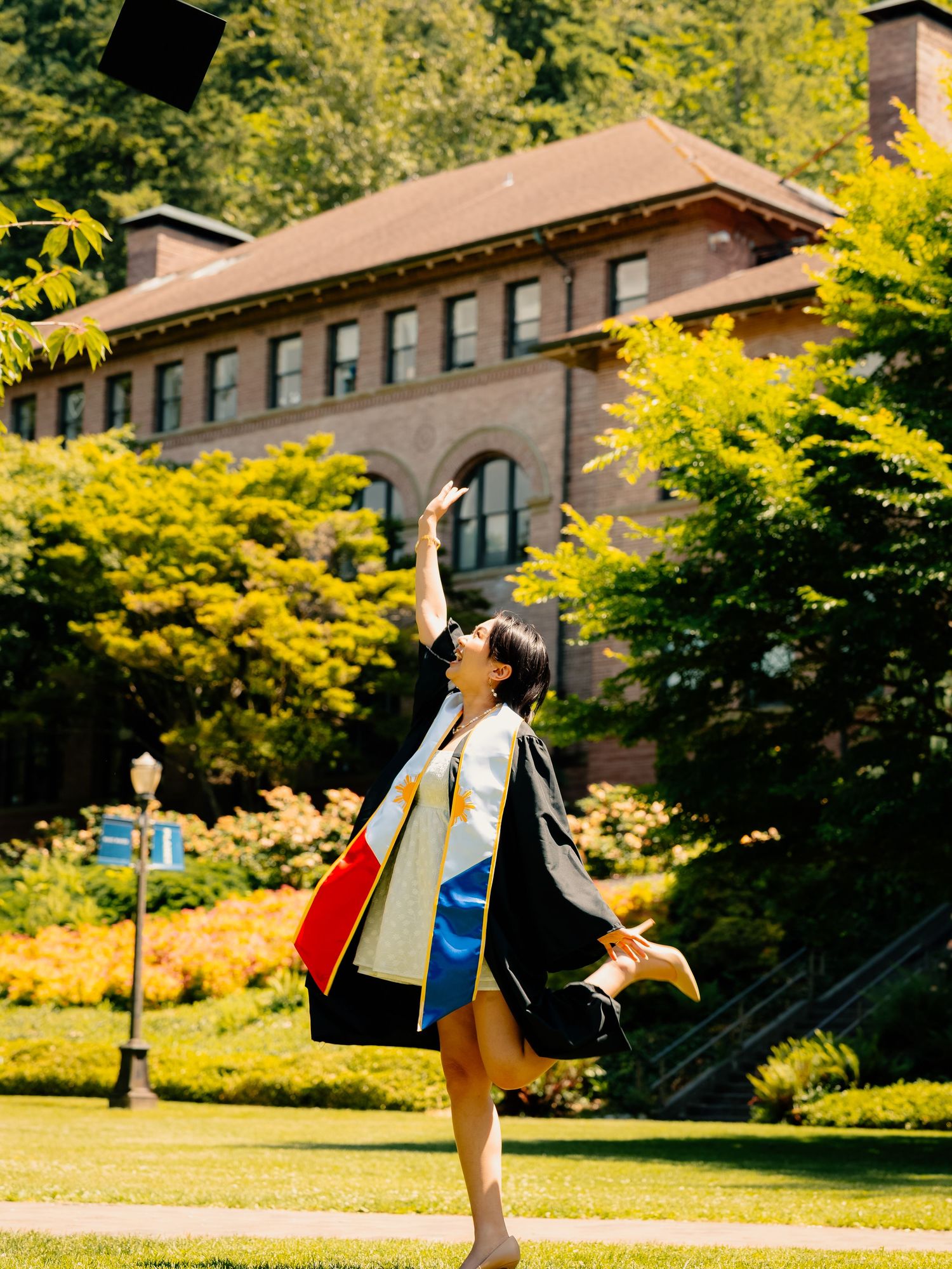 Graduate celebrates on a sunny campus lawn with yellow trees and historic brick building in the background.