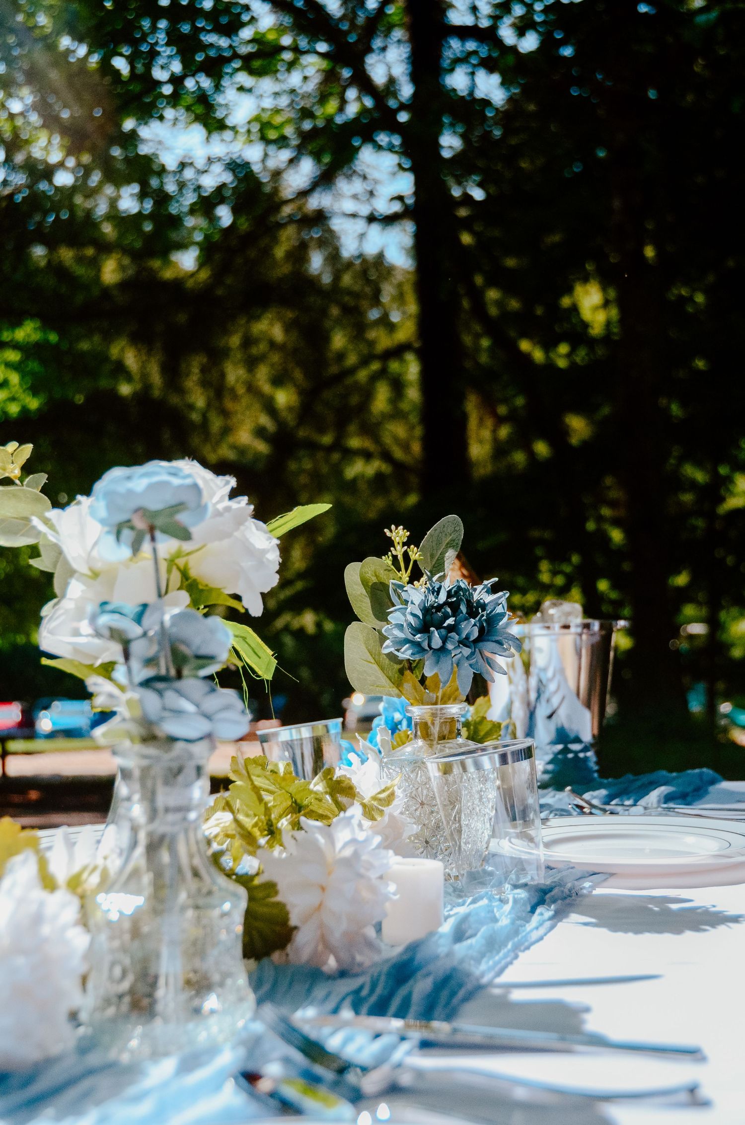 Elegant outdoor table settings with white and light blue floral centerpieces in glass vases for a garden party.