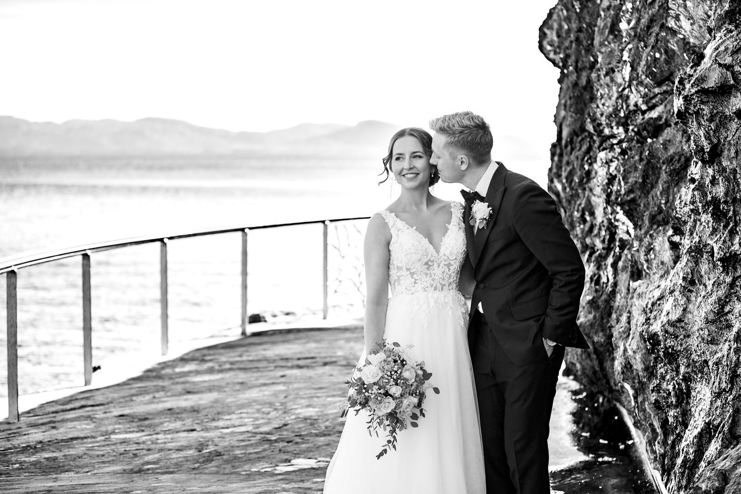 A romantic black and white wedding photo captured on a lakeside dock with mountains in the background.
