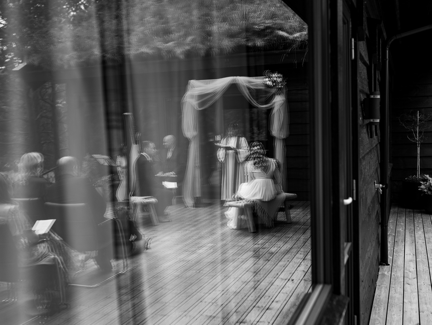 A haunting black and white photograph showing a figure hunched over on a wooden porch with mysterious reflections in glass.