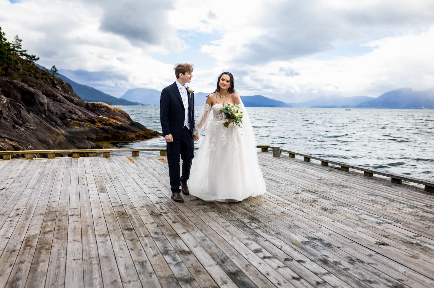 A couple stands together on a wooden pier overlooking a scenic mountain lake on their wedding day.