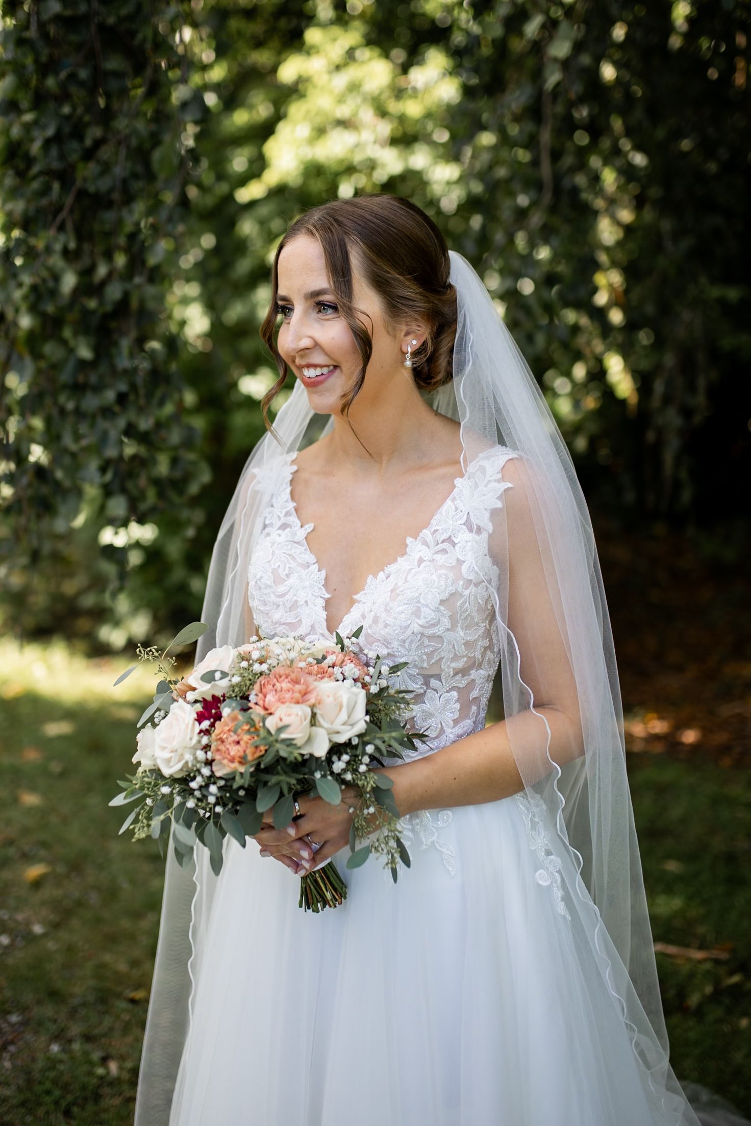 Beautiful bride in lace V-neck wedding dress and veil holds a pink and white floral bouquet in a garden setting.