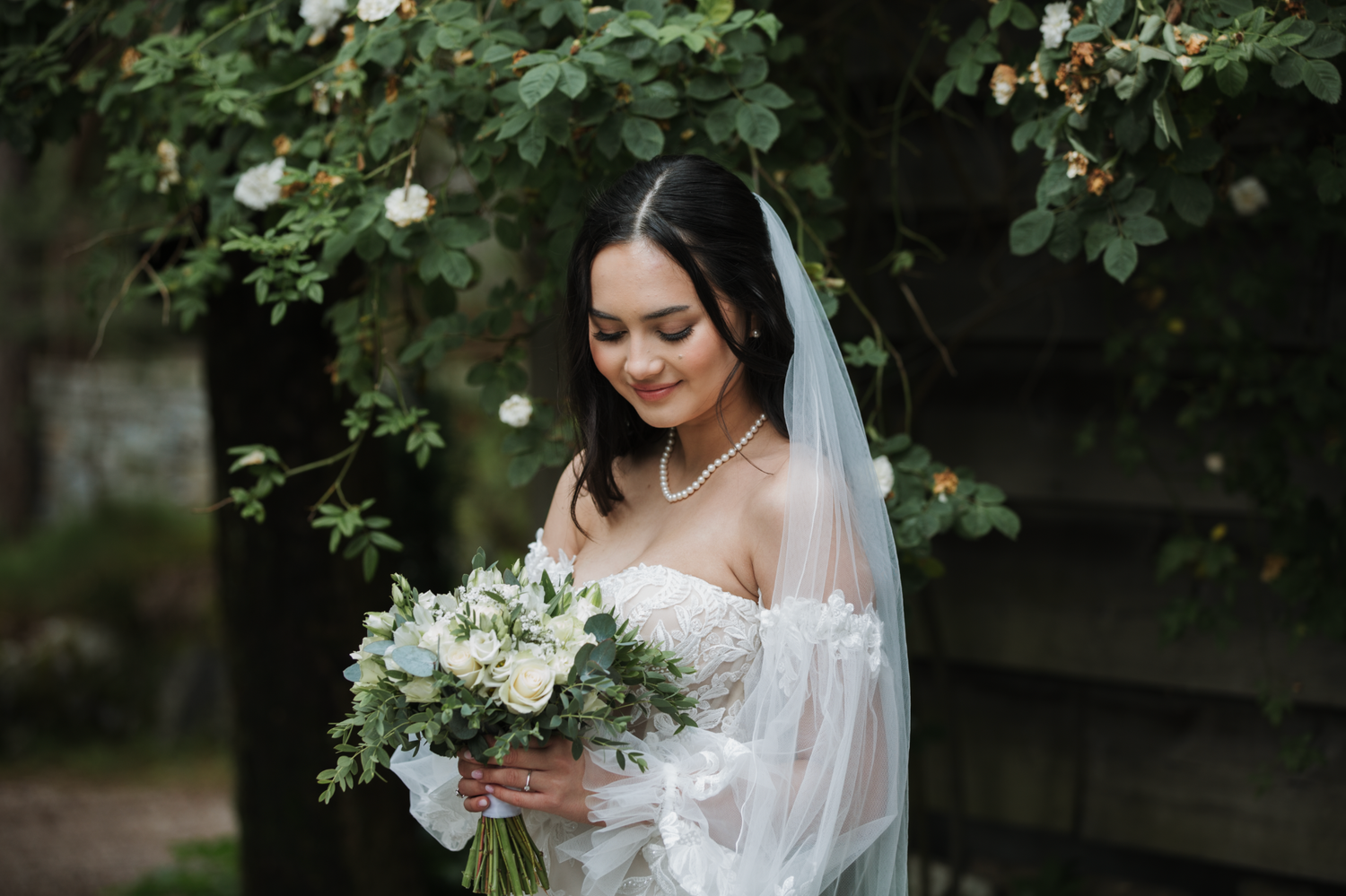 A bride in a white wedding dress holds a lush bouquet of white flowers while standing in a garden setting.