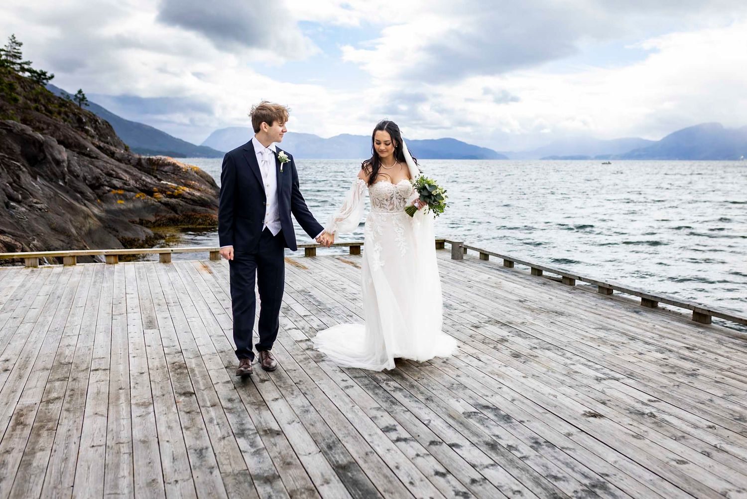 Bride and groom holding hands walking on wooden dock with scenic mountain lake backdrop on wedding day.