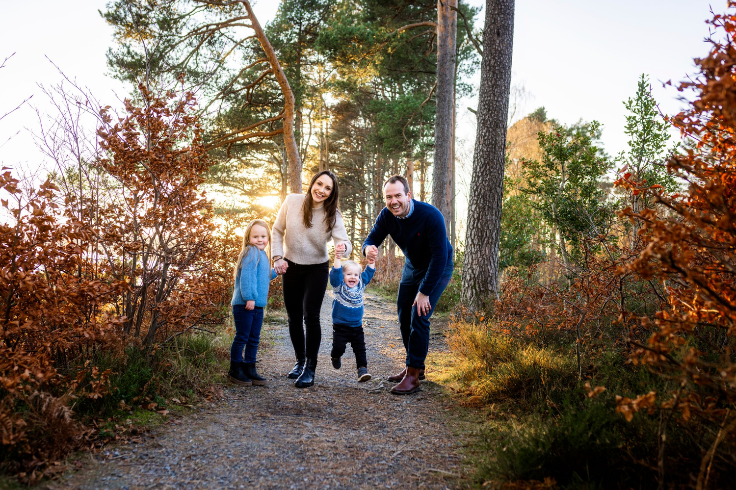 Family of four smiling on a forest path surrounded by autumn foliage with golden sunset light filtering through pine trees.
