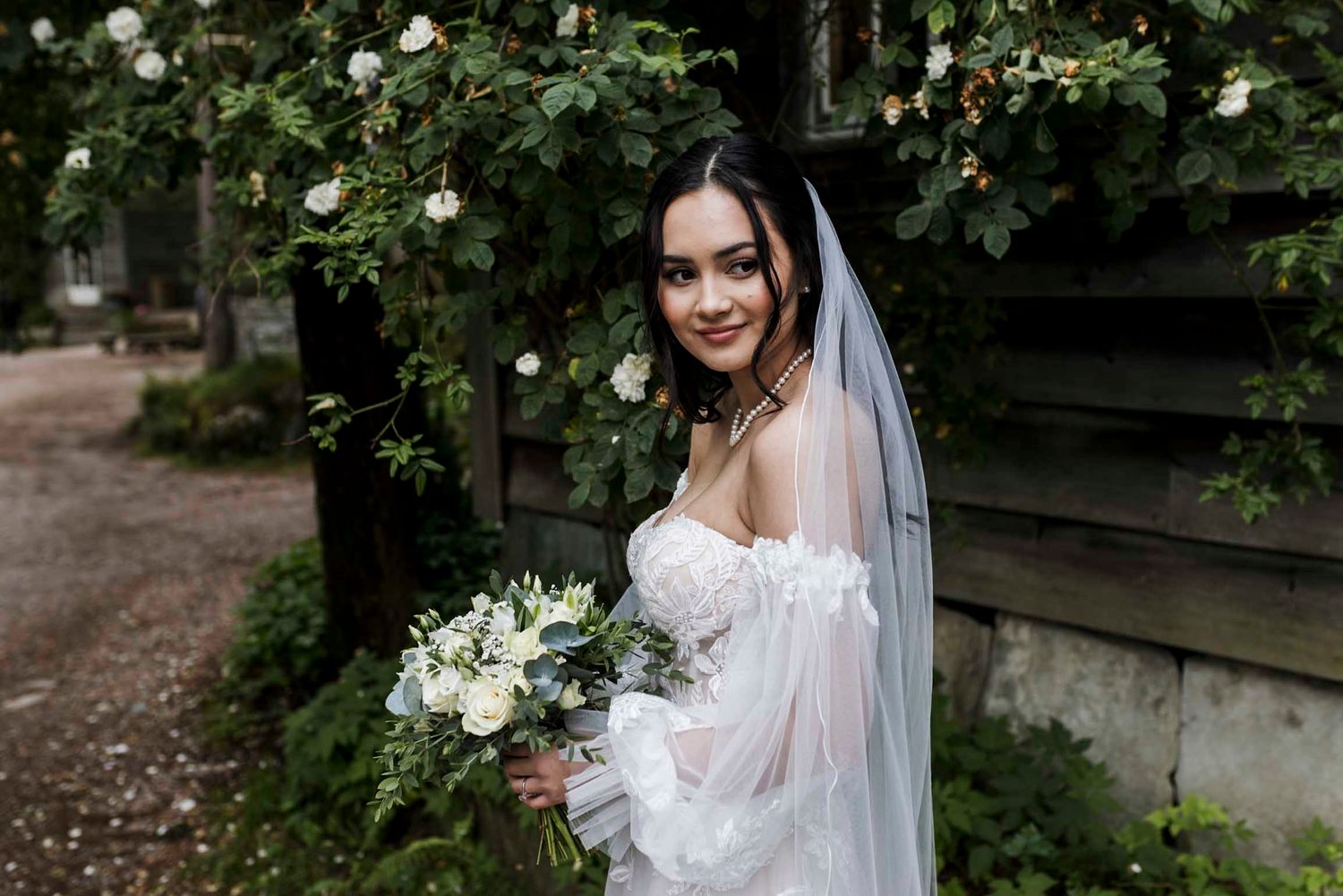 Bride in white off-shoulder gown and veil holds floral bouquet, posing outdoors near lush greenery.