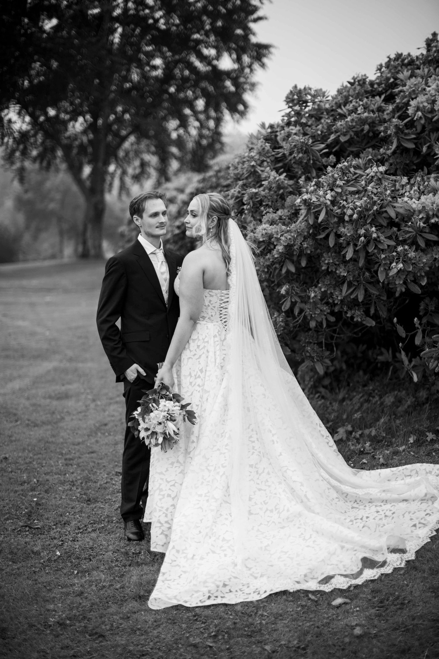Elegant bride and groom pose near blooming shrubs in black and white wedding portrait outdoors.