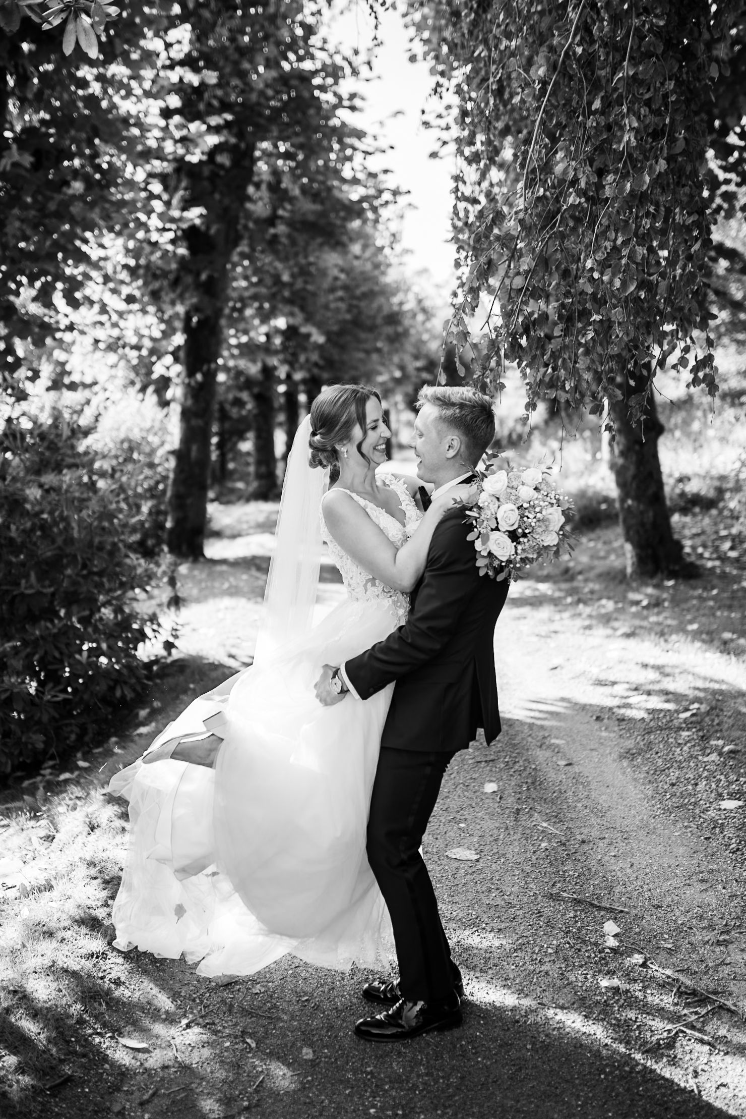 Newlyweds embrace and dance on a tree-lined path during their romantic black and white wedding photoshoot.