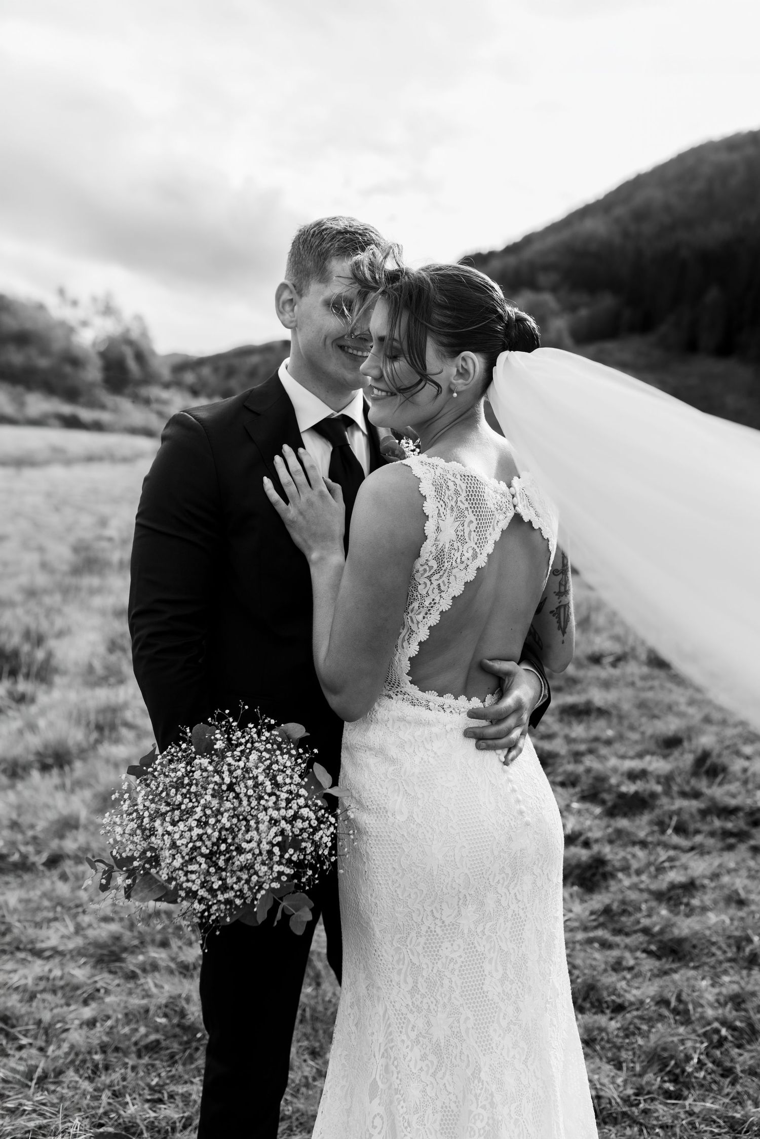 Black and white photo of a bride and groom embracing outdoors, her veil flowing, holding a bouquet in a scenic field.