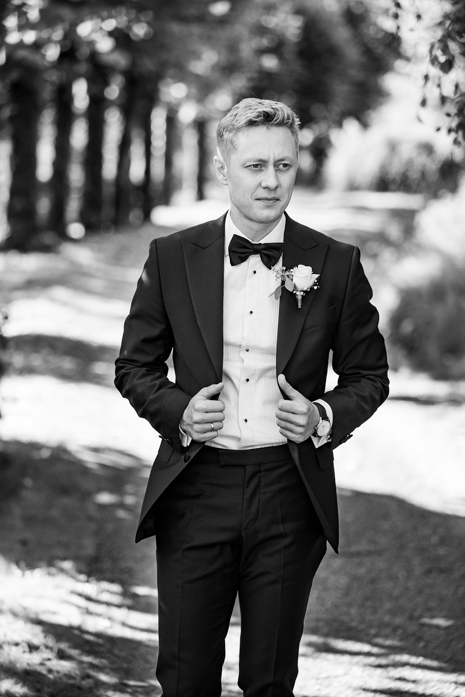 Black and white portrait of a groom in classic tuxedo with bowtie and boutonniere standing outdoors on a wooded path.