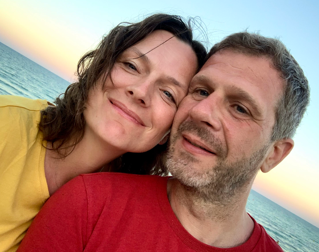 A couple takes a smiling selfie together at the beach during sunset, with one wearing yellow and one in red.