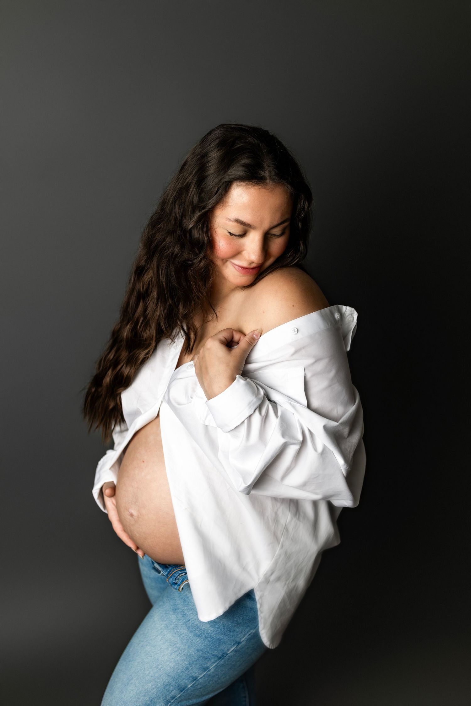 Smiling pregnant woman in white off-shoulder shirt and jeans cradling her bare baby bump against a dark gray background.
