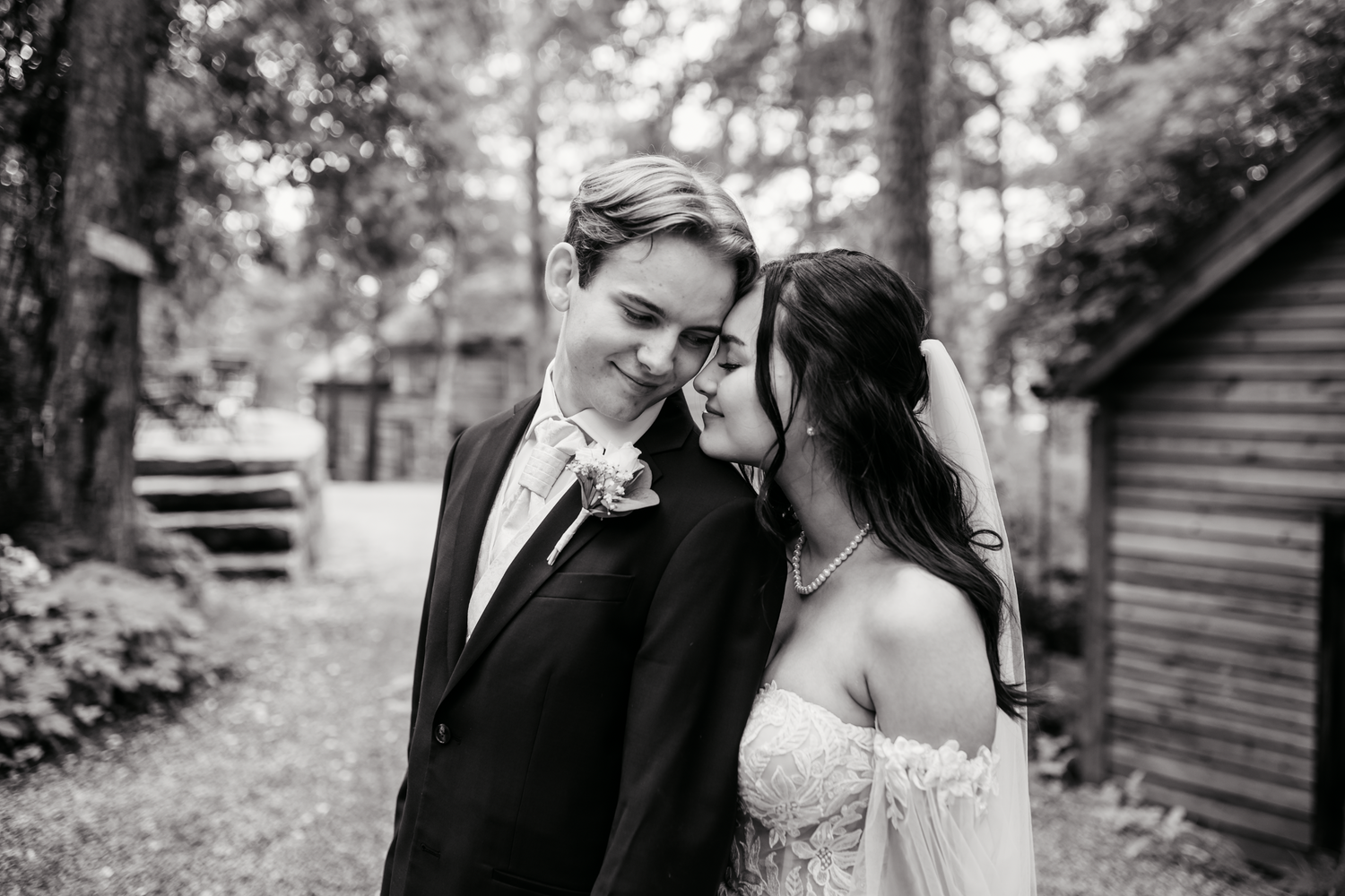 A black and white wedding portrait of a couple sharing an intimate moment in front of a rustic cabin in the woods.